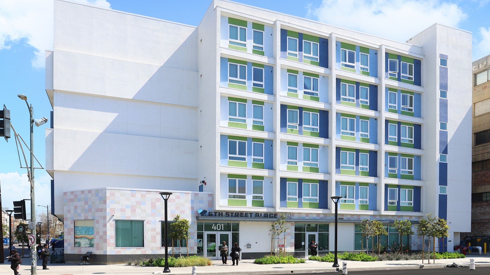 A modern five-story apartment building with white walls, green and blue window accents, offers affordable housing at 5th Street Place, 401. A few people are walking nearby on the sidewalk.