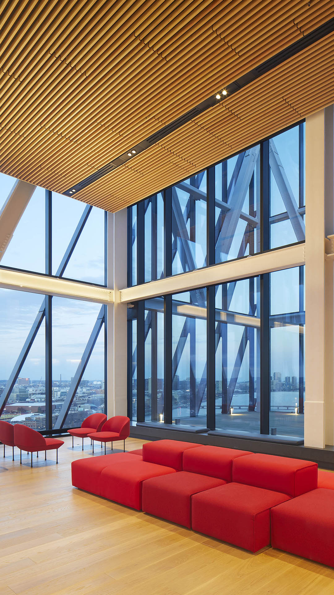 A modern lounge designed for higher education spaces, featuring red modular seating and armchairs, wood flooring and ceiling, plus large floor-to-ceiling windows with steel beams framing a stunning cityscape.