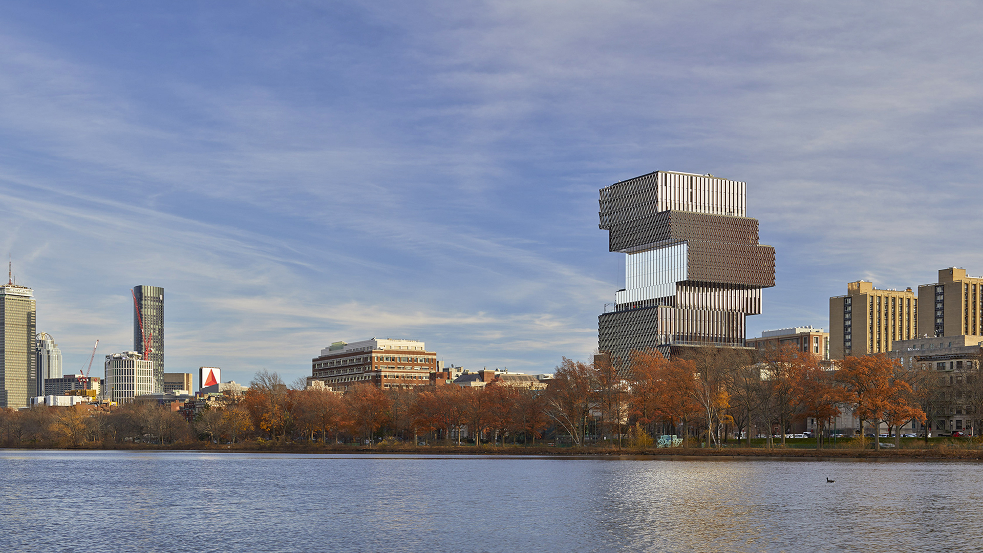 A city skyline with modern and historic buildings, including a uniquely stacked, block-shaped structure—home to a renowned higher education institution—seen across a river lined with autumn trees under a blue sky.