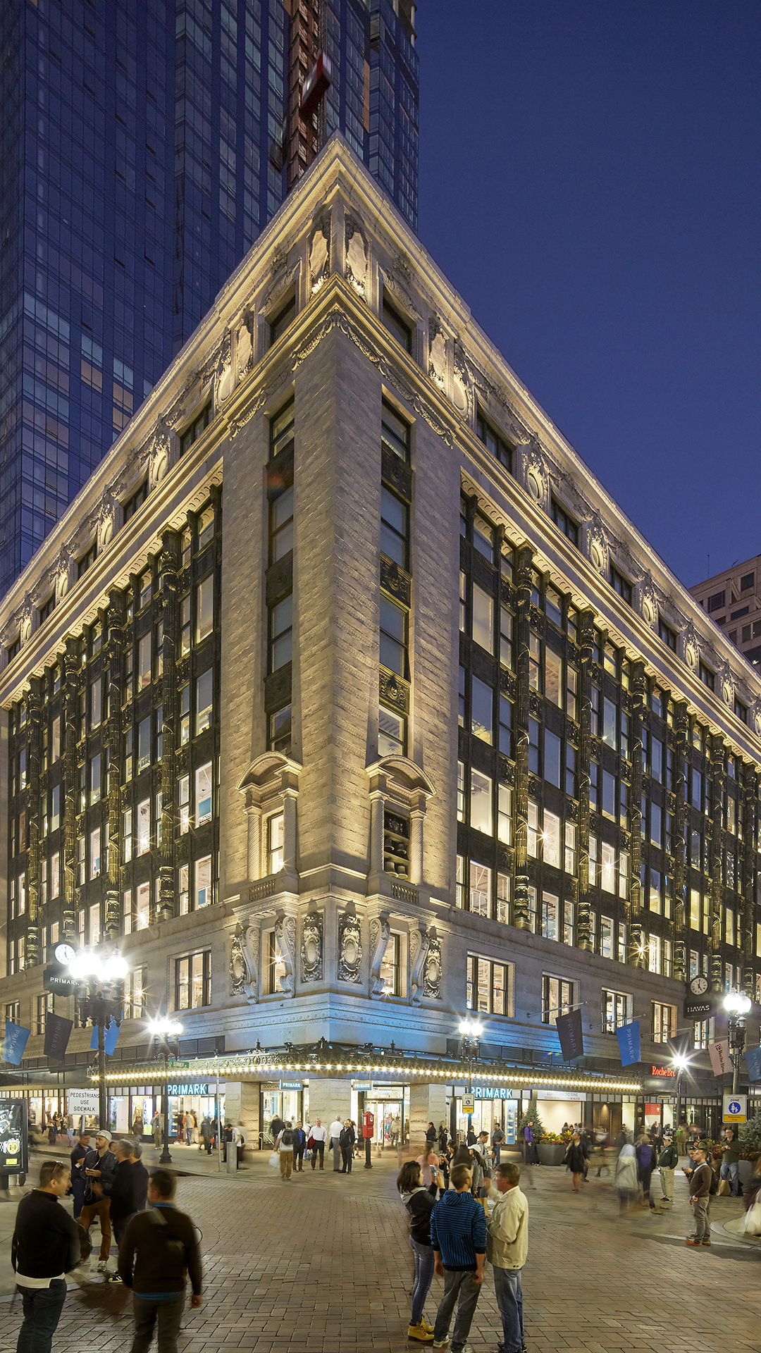 A historic, multi-story retail department store building with ornate architecture and bright lights stands on a busy city street corner at dusk. People gather near the entrance under a blue awning as evening falls.