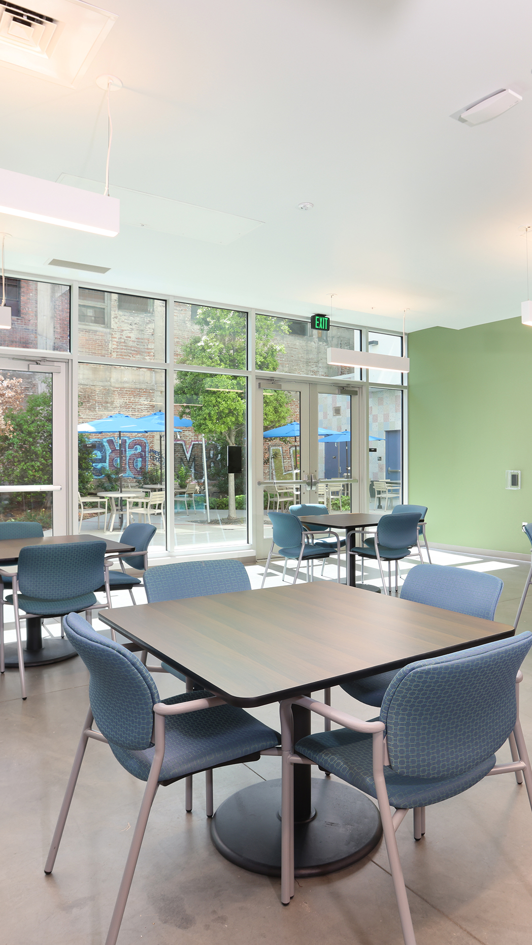 A modern, bright dining area with empty tables and blue chairs, large windows letting in natural light, and a view of outdoor seating and greenery in an affordable housing community.
