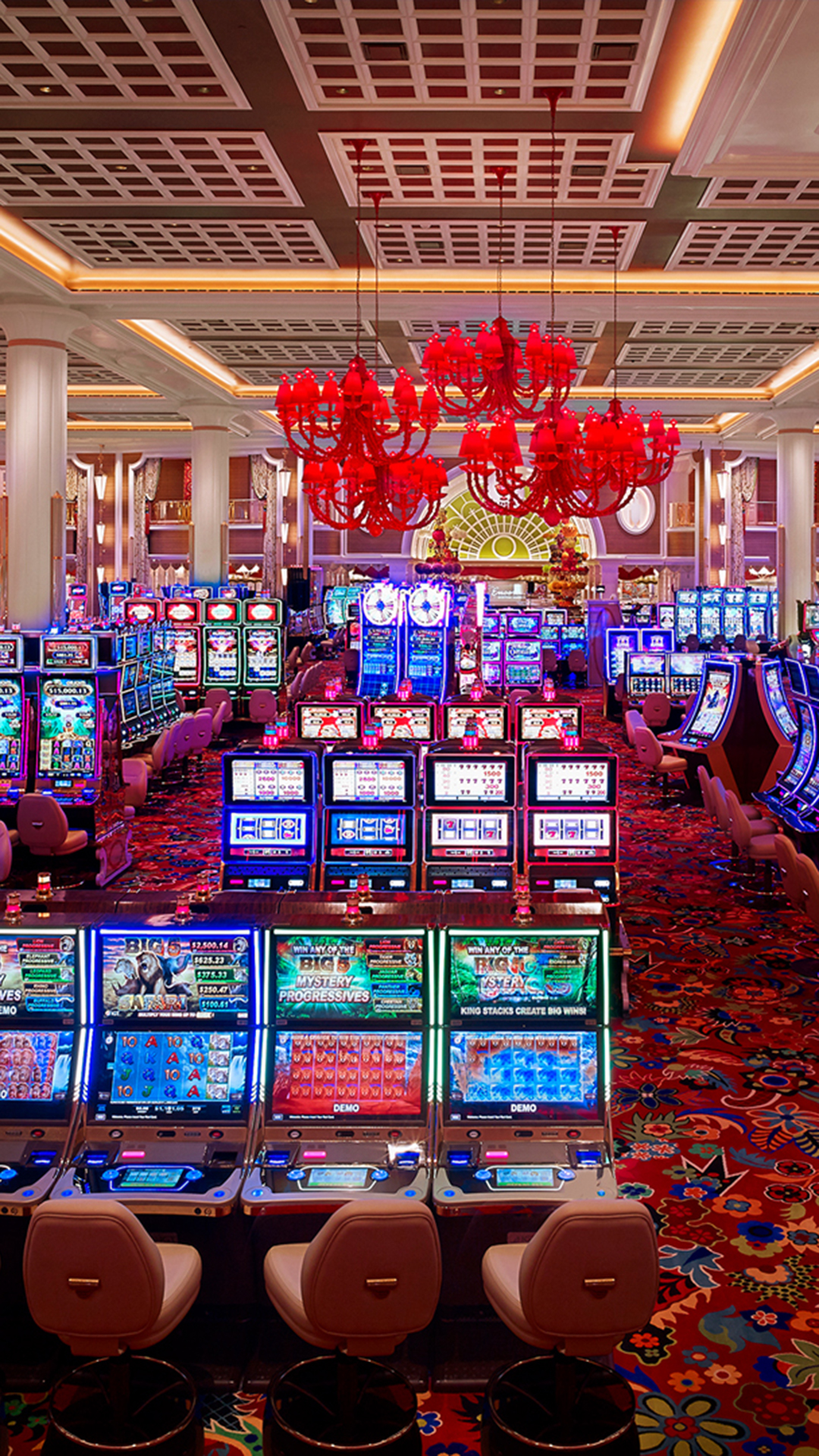 A brightly lit casino interior features rows of gaming slot machines with colorful displays and empty chairs, red chandeliers, patterned carpet, and a lively, luxurious atmosphere.