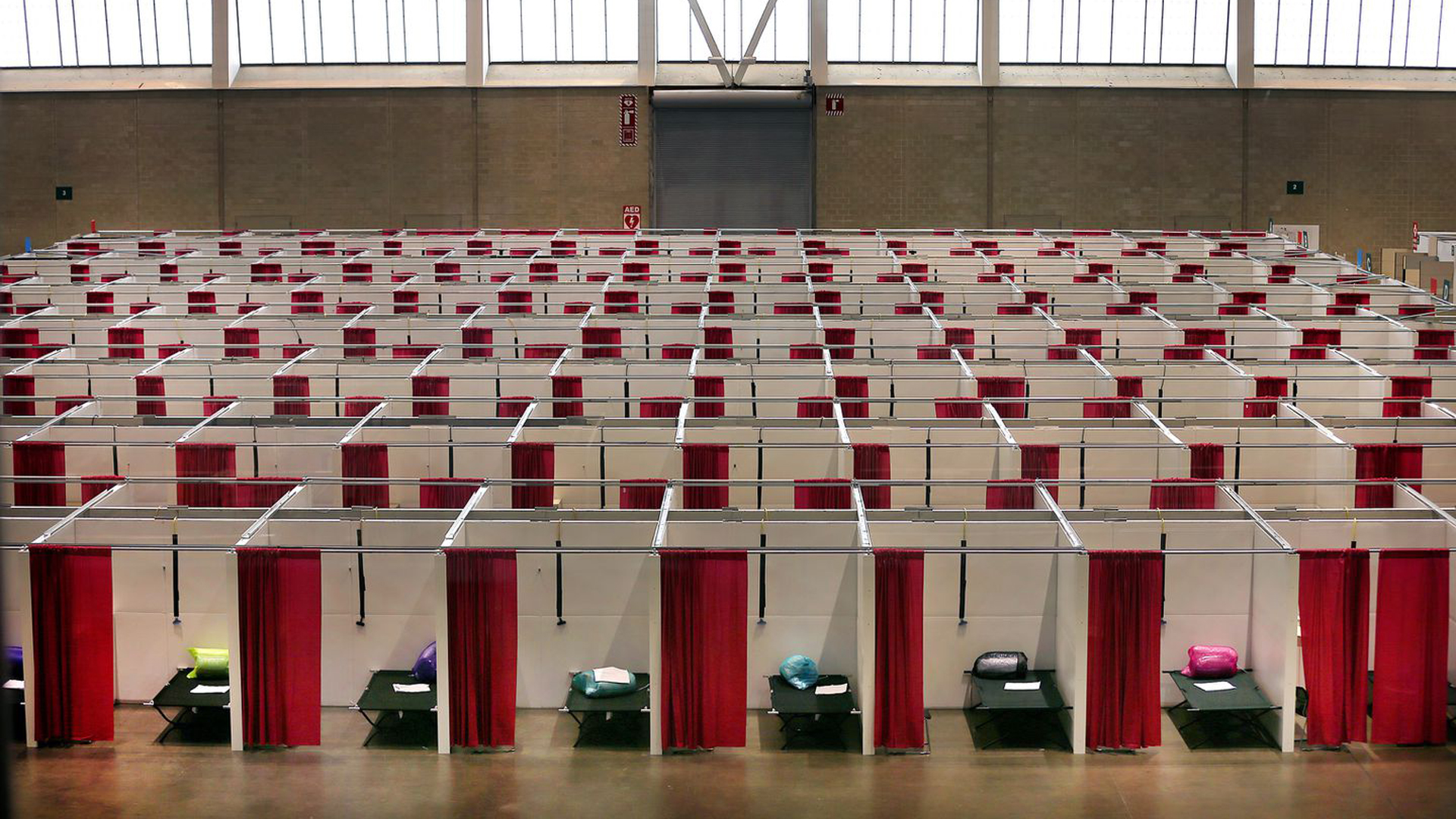 Rows of small makeshift cubicles with red curtains and cots inside, set up in a large indoor facility with high windows, resemble a temporary shelter or field hospital organized by the Federal Government.
