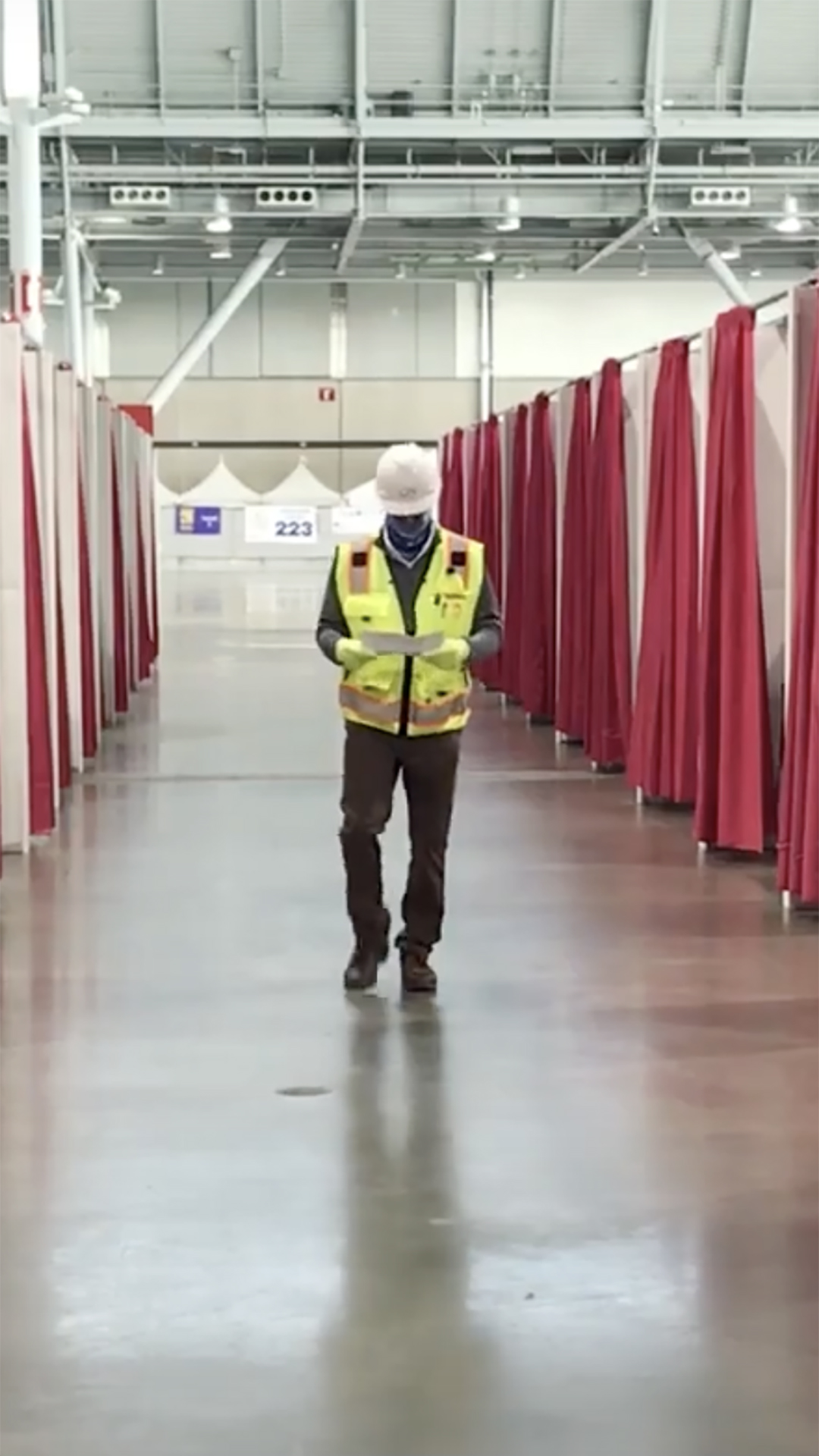A person wearing a yellow safety vest and white hard hat, possibly working for the Federal Government, walks down a hallway lined with red and white curtains in an indoor facility.
