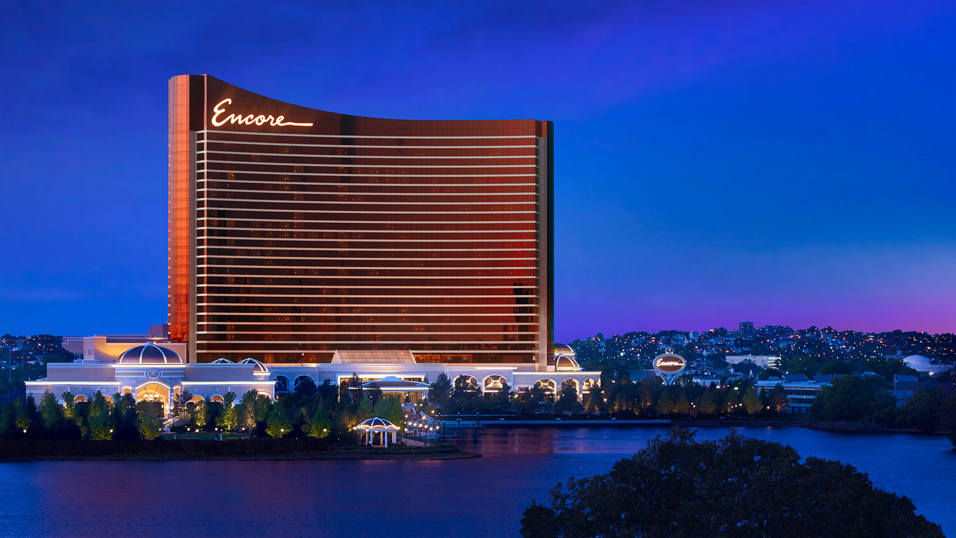 A tall, modern hotel building with Encore illuminated at the top stands by the water at dusk, its exterior glowing with lights against a deep blue and purple sky—an inviting hub for luxury stays and gaming. Trees and smaller lit buildings surround the hotel.
