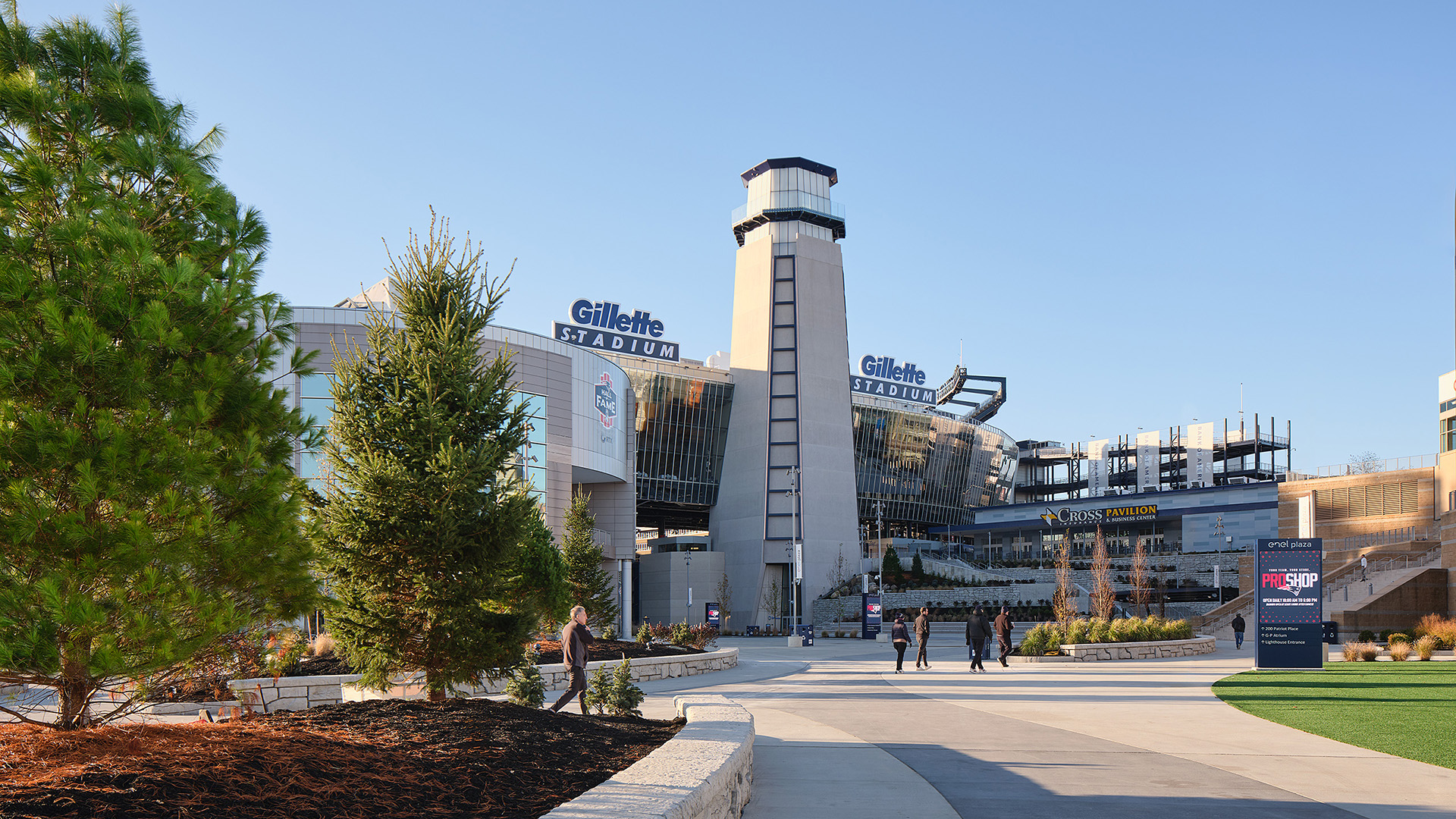 A wide view of Gillette Stadium’s entrance, featuring a tall tower, “Gillette Stadium” signs, and a few people walking along a curved pathway bordered by trees on a clear day—an iconic destination for sports and entertainment.
