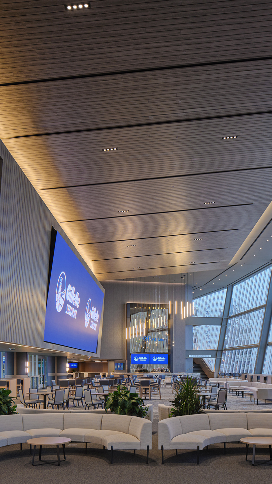 A spacious, modern entertainment lounge with curved white sofas, tables, large plants, and high ceilings. A giant screen displays “Gillette Stadium.” Floor-to-ceiling windows fill the space with natural light and contemporary lights hang overhead.
