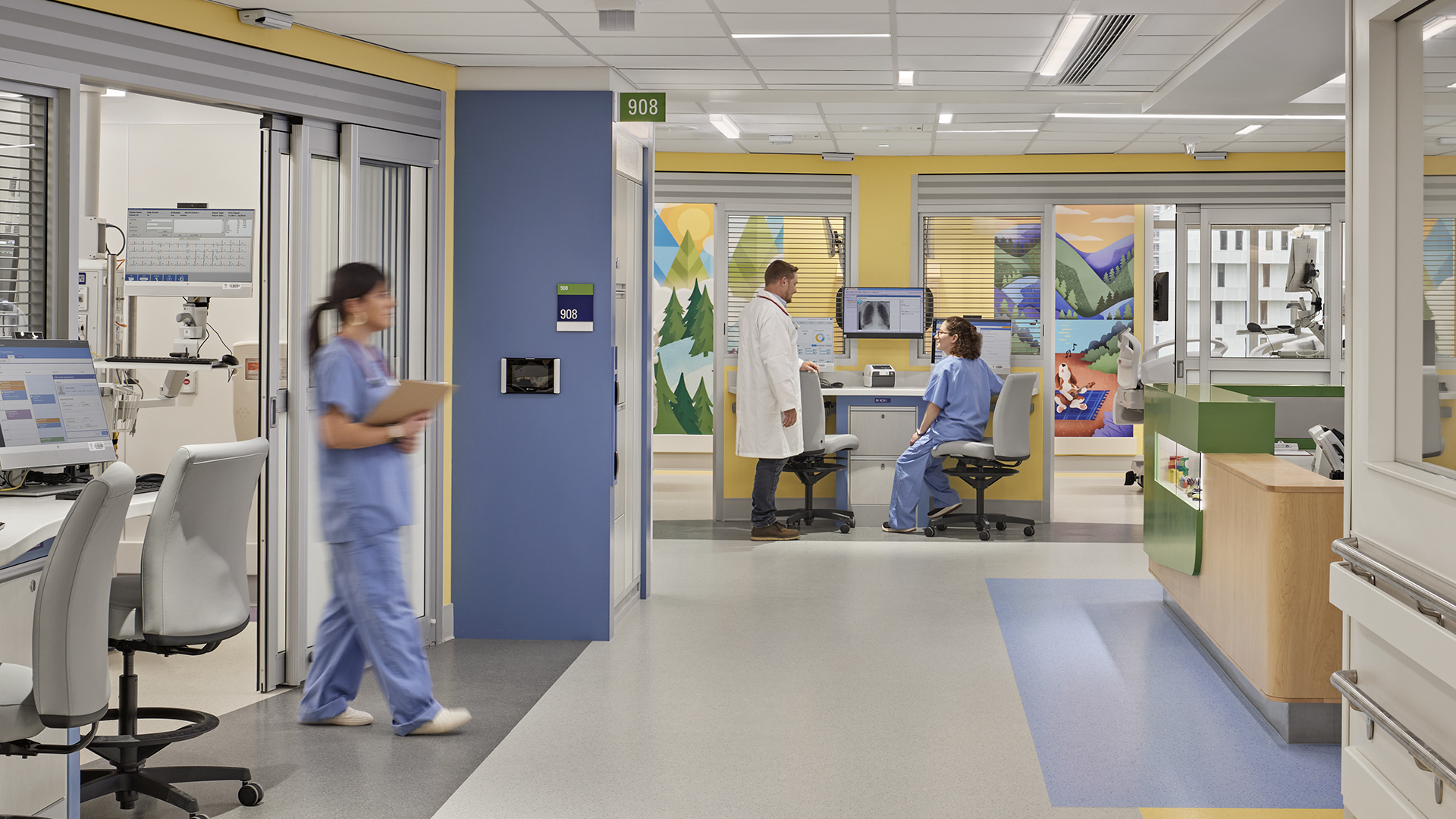 A doctor speaks with a seated patient in a colorful, modern hospital room. A nurse in blue scrubs walks by, while another works at a desk with computers in the foreground. The clean, bright environment reflects high-quality healthcare.