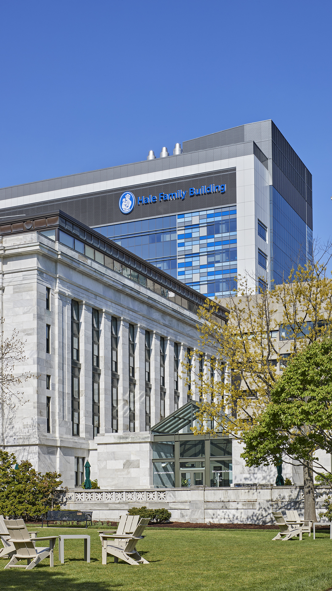 A modern and historic healthcare building, the Hale Family Building features white stone columns and glass windows, standing under a clear blue sky with trees and lawn chairs in the foreground.