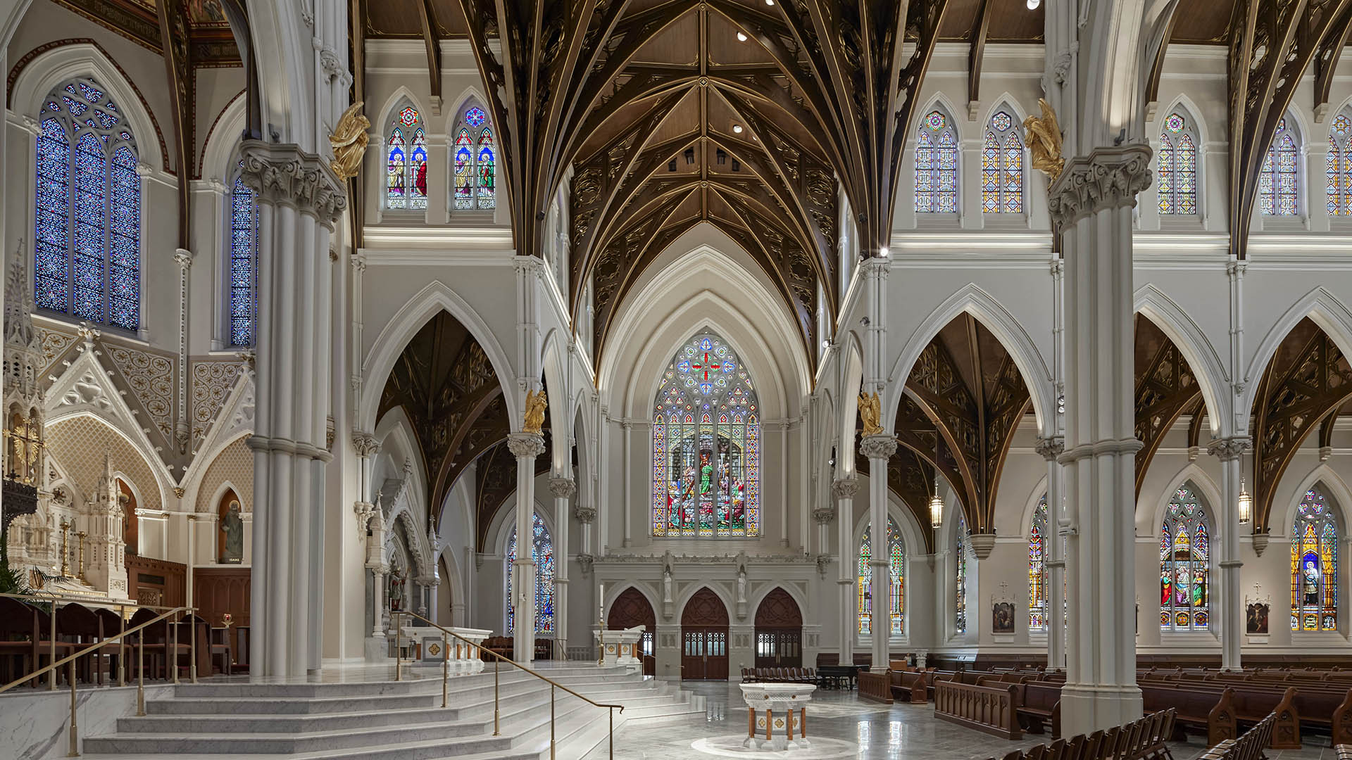 Interior of a grand nonprofit cathedral with high vaulted ceilings, ornate columns, large stained glass windows, and detailed arches. Sunlight streams through colorful glass, illuminating the altar and pews below.