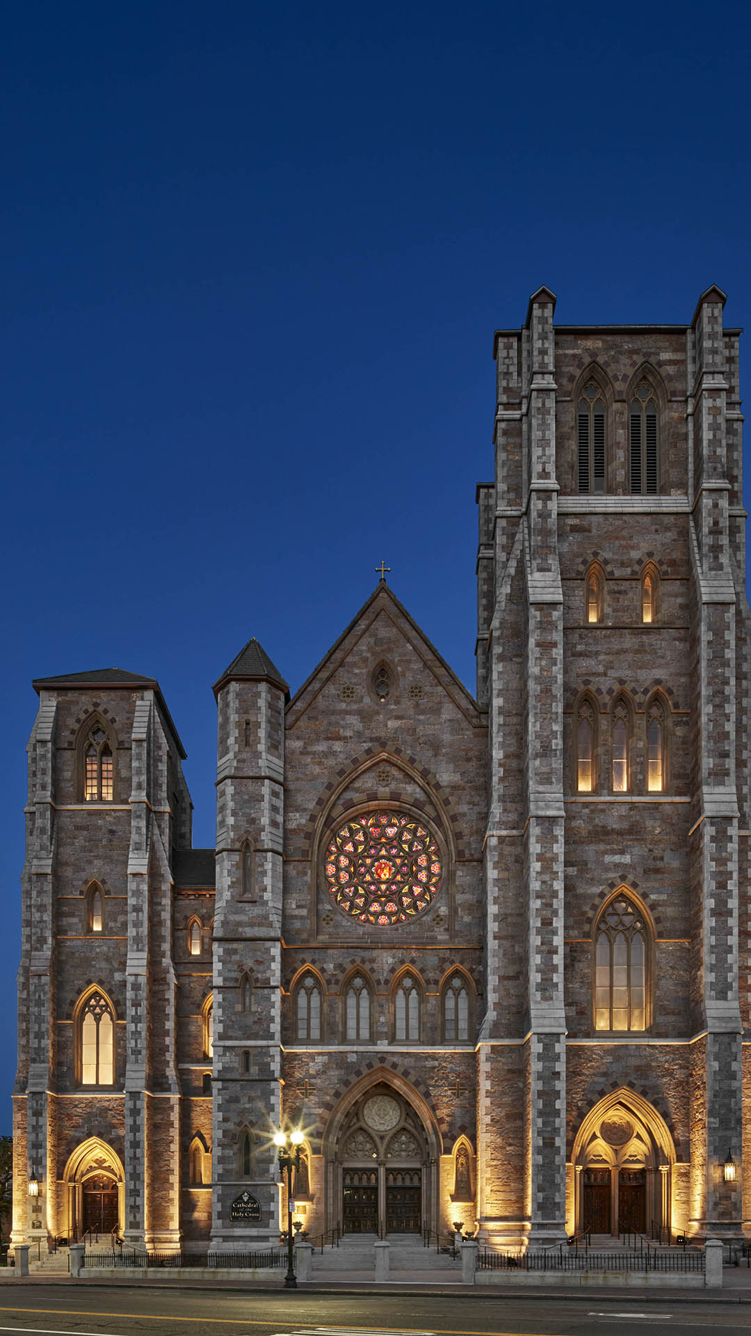 The image shows a large Gothic-style cathedral, managed by a local nonprofit, with two tall towers and a central rose window, illuminated by warm lights against a deep blue evening sky.