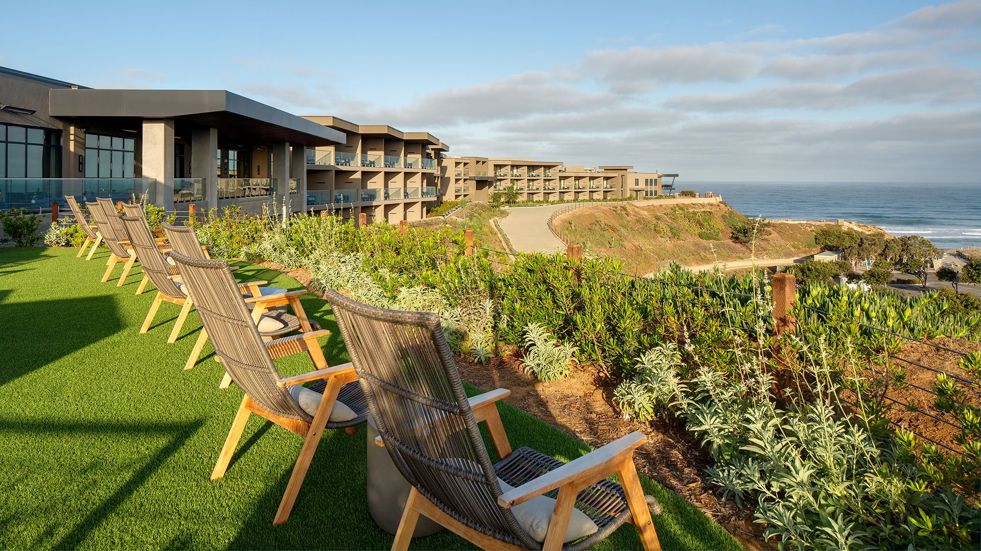 Wooden lounge chairs are arranged on a grassy terrace overlooking an oceanfront resort, showcasing the hospitality of lush landscaping and buildings set on a coastal bluff under a partly cloudy sky.