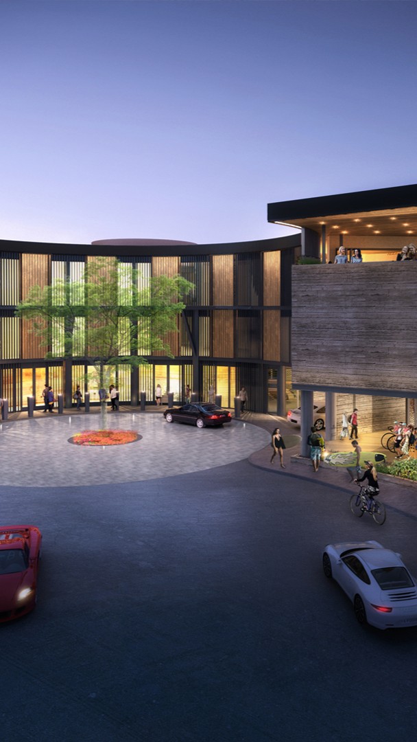 A modern hospitality building with large windows and wooden accents surrounds a circular driveway featuring a small tree. People walk, bike, and gather outside while cars pass by at dusk.