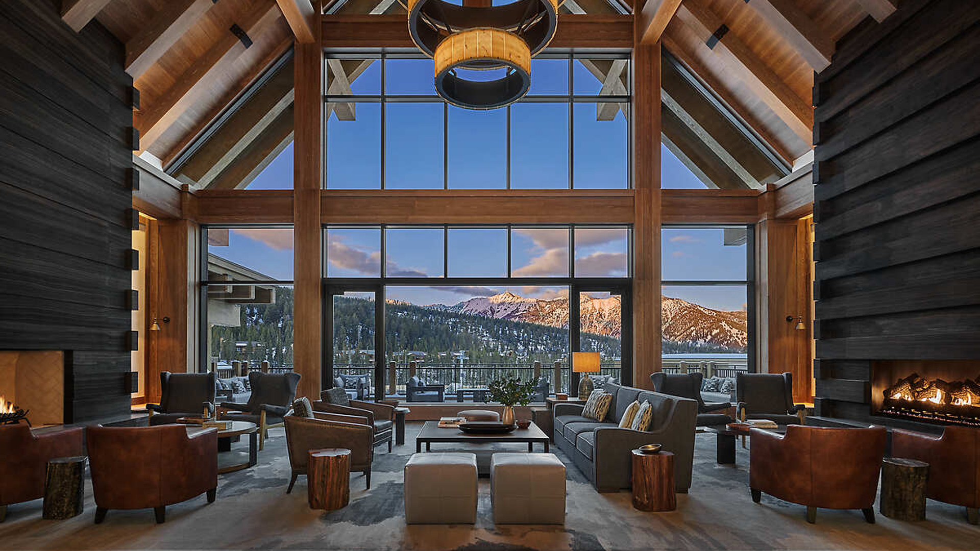 A luxurious lodge living room with high wood-beamed ceilings, large windows showcasing snowy mountains, cozy seating, a modern fireplace, and warm ambient lighting—perfect for experiencing elevated hospitality.