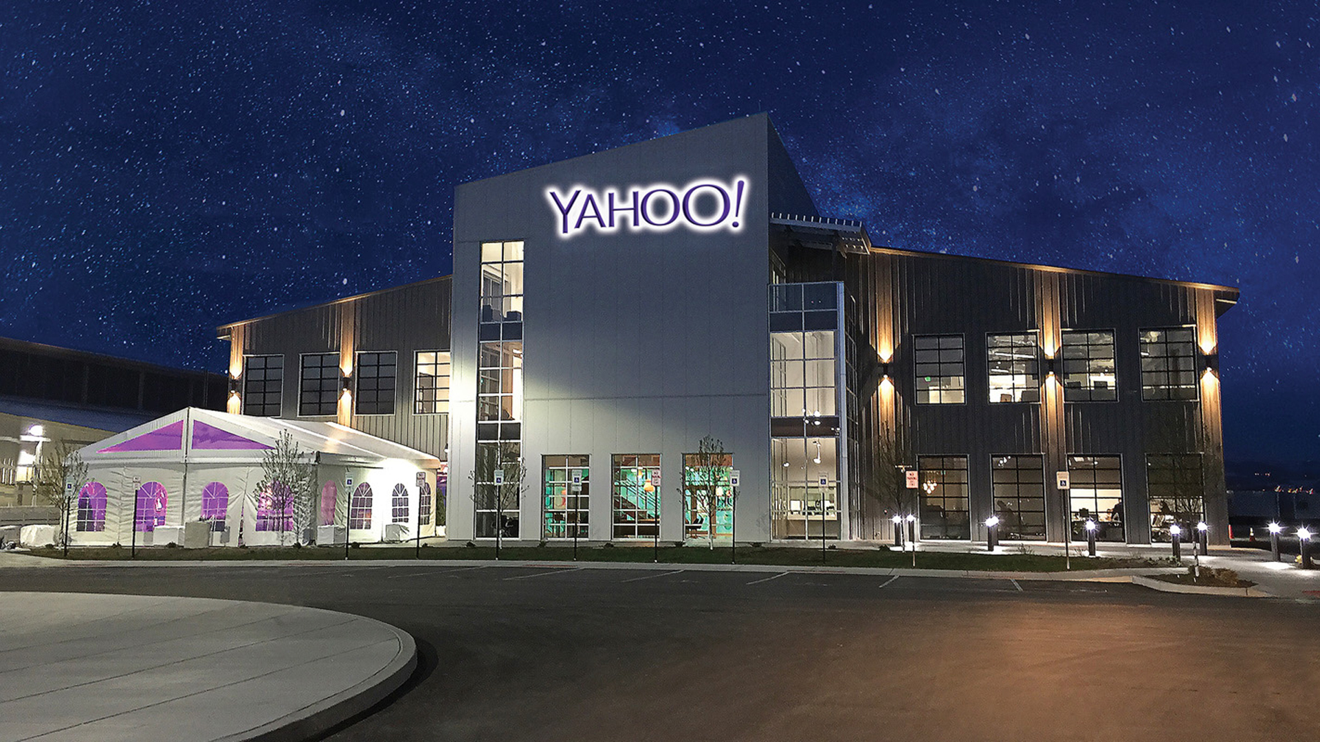 A modern, mission critical building with large windows and a prominent “YAHOO!” sign is illuminated at night under a starry sky. A white tent with purple lighting stands to the left, while the parking lot in front is mostly empty.