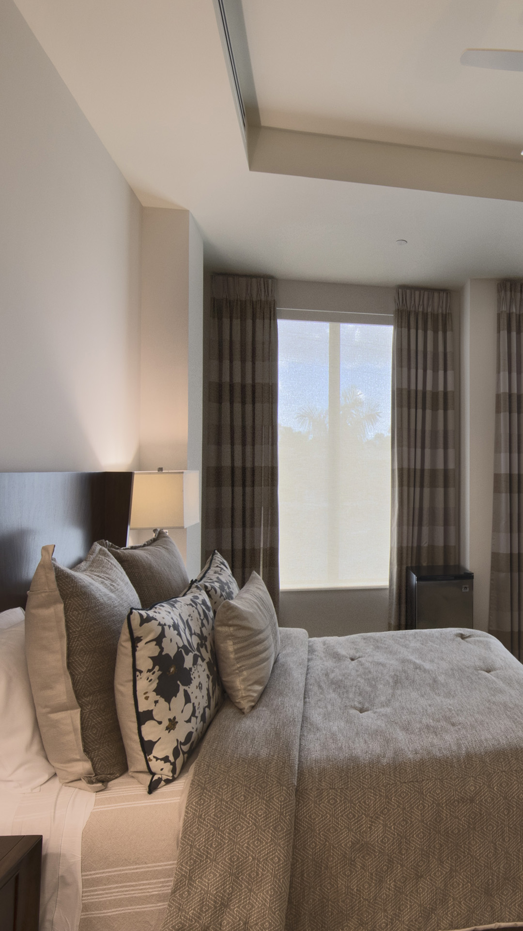 A neatly made bed with gray and patterned pillows sits beside a dark nightstand and lamp in this inviting assisted living room, featuring beige walls, checkered curtains, and a large window with a view of trees outside.