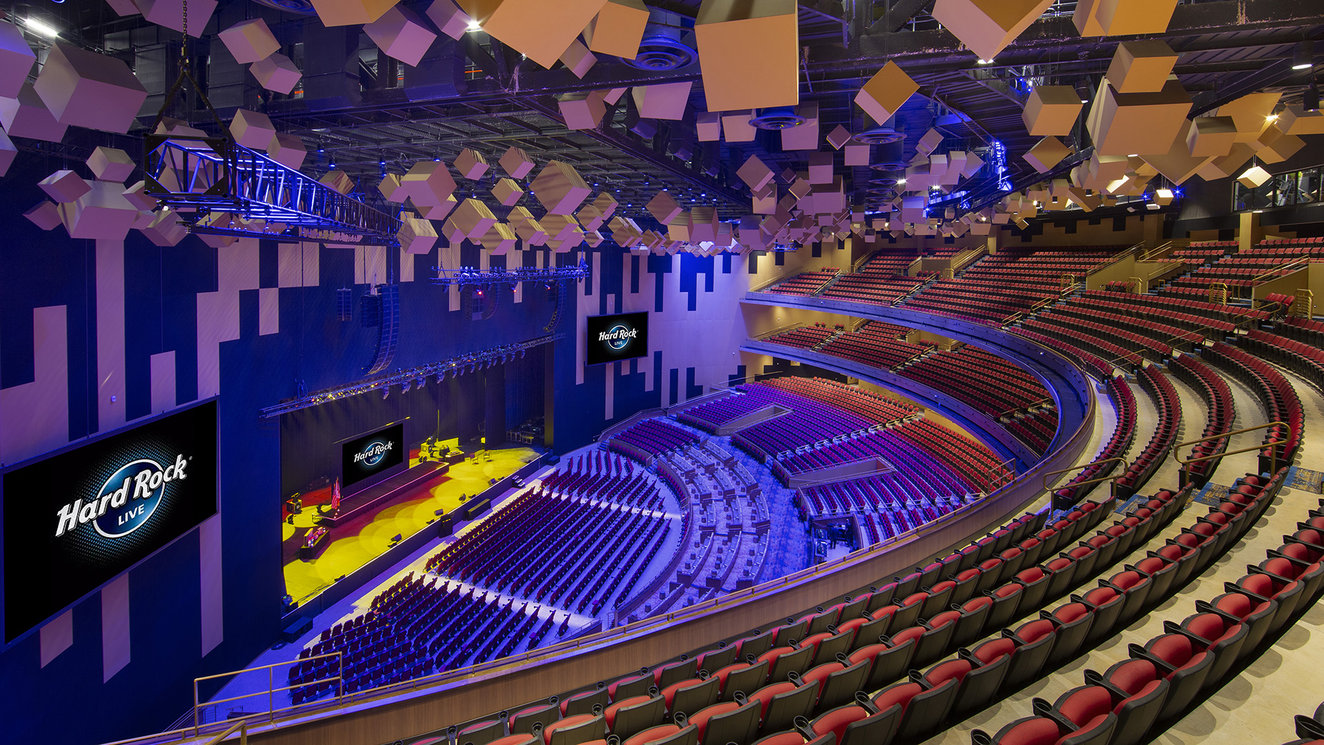 Spacious, modern concert hall designed for top-tier entertainment, featuring curved rows of red seats, a large stage with Hard Rock Live screens, and geometric acoustic panels. Blue and yellow lighting highlights the empty venue.