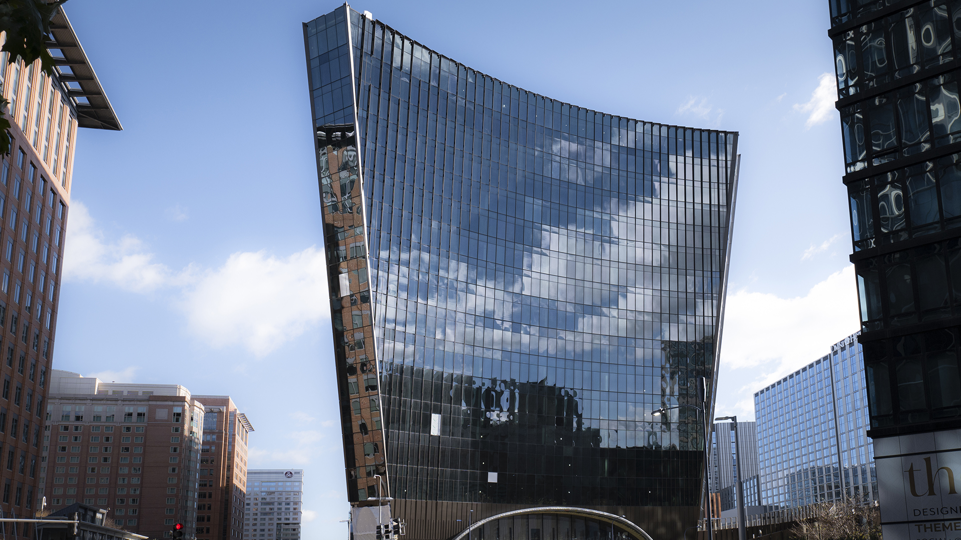 A modern glass skyscraper with a distinctive curved design, housing leading Life Sciences firms, reflects the blue sky and clouds, surrounded by other tall city buildings on a sunny day.