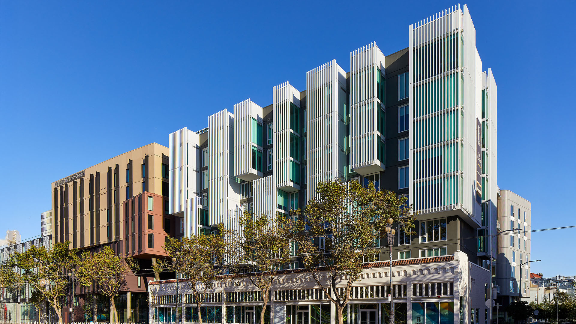 Modern residential multi-story building with prominent vertical metal fins and glass windows, featuring geometric designs. Trees line the street in front, and the sky is clear and blue.