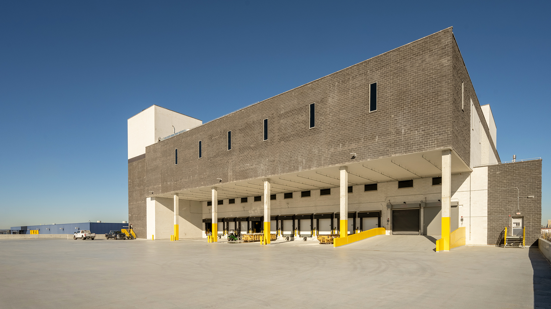 A large, modern industrial warehouse with a gray facade, multiple distribution loading docks, yellow safety barriers, and a wide concrete lot under a clear blue sky.