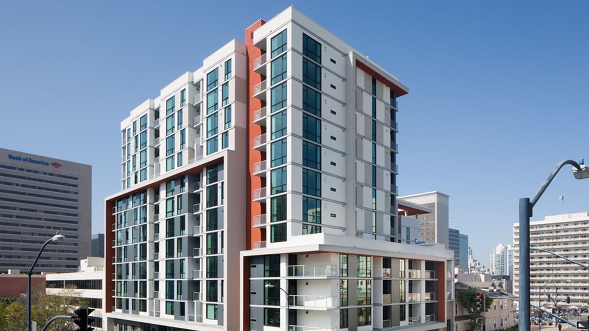 A modern mid-rise apartment building with large glass windows and balconies, set against a clear blue sky and surrounded by other city buildings, offers stylish affordable housing in an urban setting.