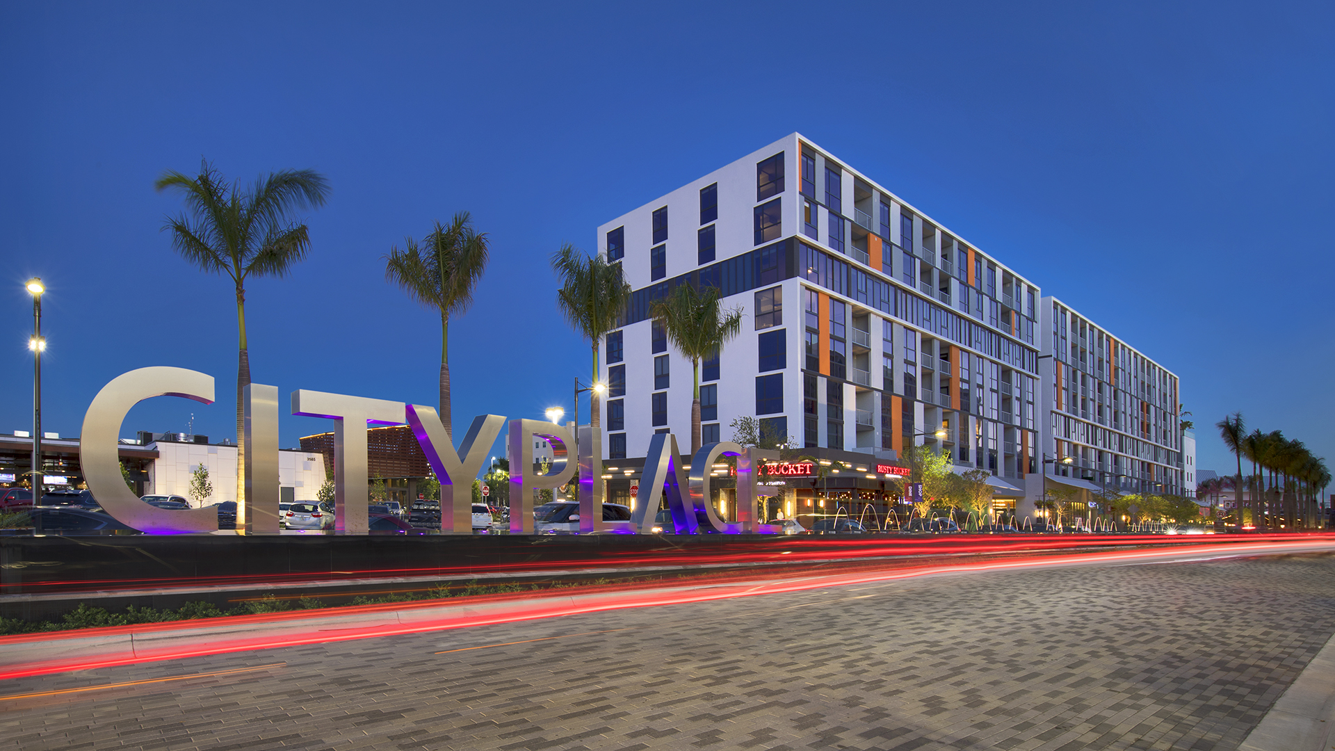 Modern building at dusk with large CITYPLACE sign in front, palm trees lining the street, and streaks of car lights passing by on a paved road. The building features many windows and retail options, including a restaurant at its base.