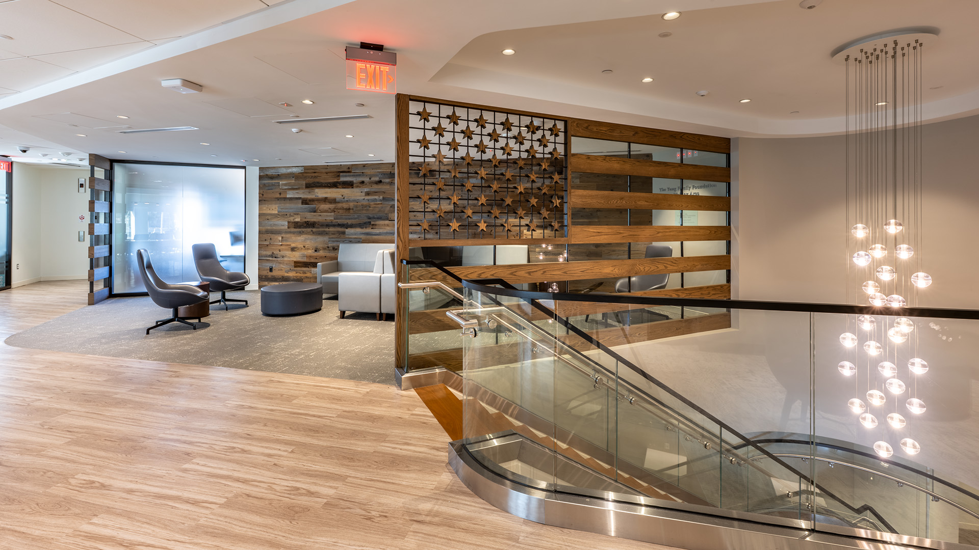 Modern nonprofit office lobby with wooden floors, a glass staircase, stylish chairs and tables, a decorative wooden divider, and a cluster of hanging pendant lights. A frosted glass wall and ceiling lights enhance the contemporary decor.