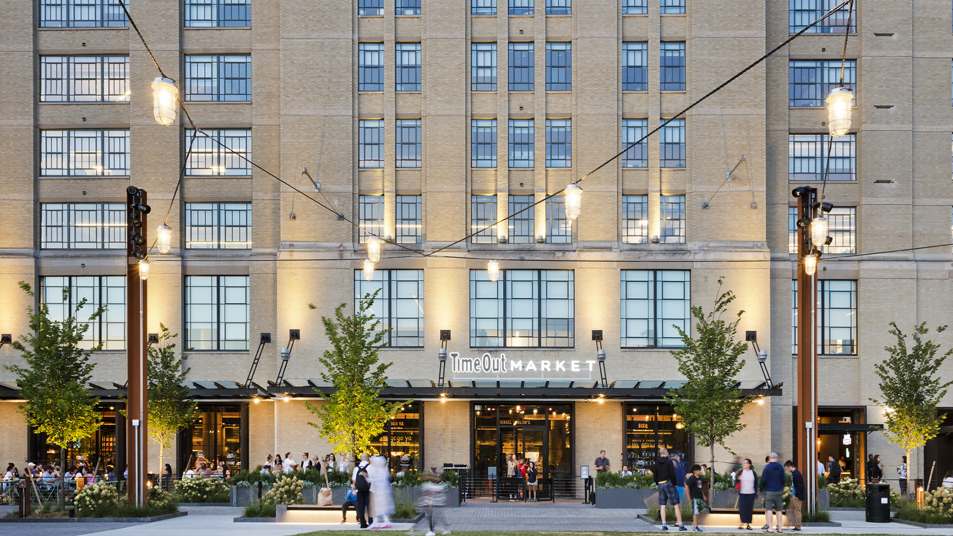People gather outside the entrance of Time Out Market, a vibrant retail hub housed in a large brick building with rows of windows and string lights overhead. Trees and outdoor seating are visible in the foreground during daylight.