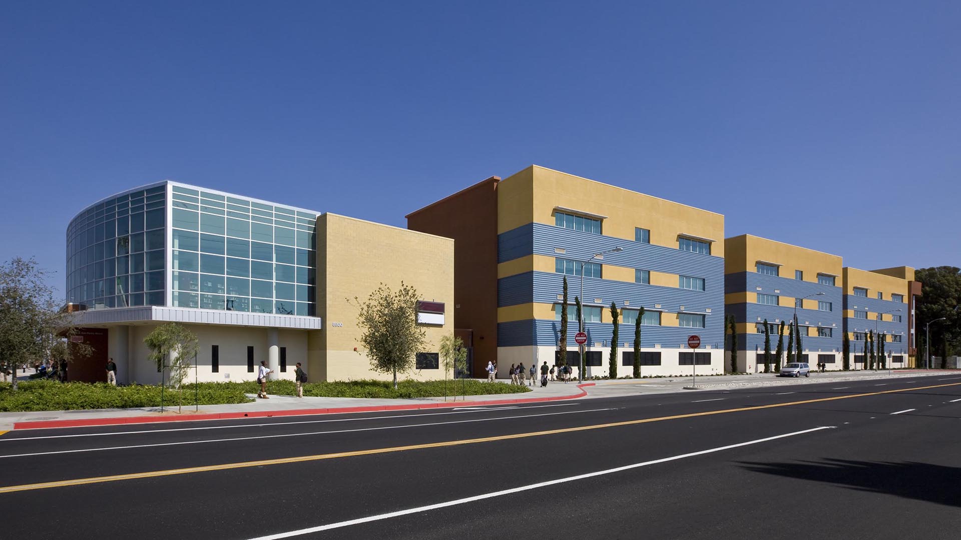 A modern K-12 school building with yellow and blue exterior walls, large windows, and a distinctive glass-fronted section. People walk on the sidewalk beside a tree-lined street under clear skies.