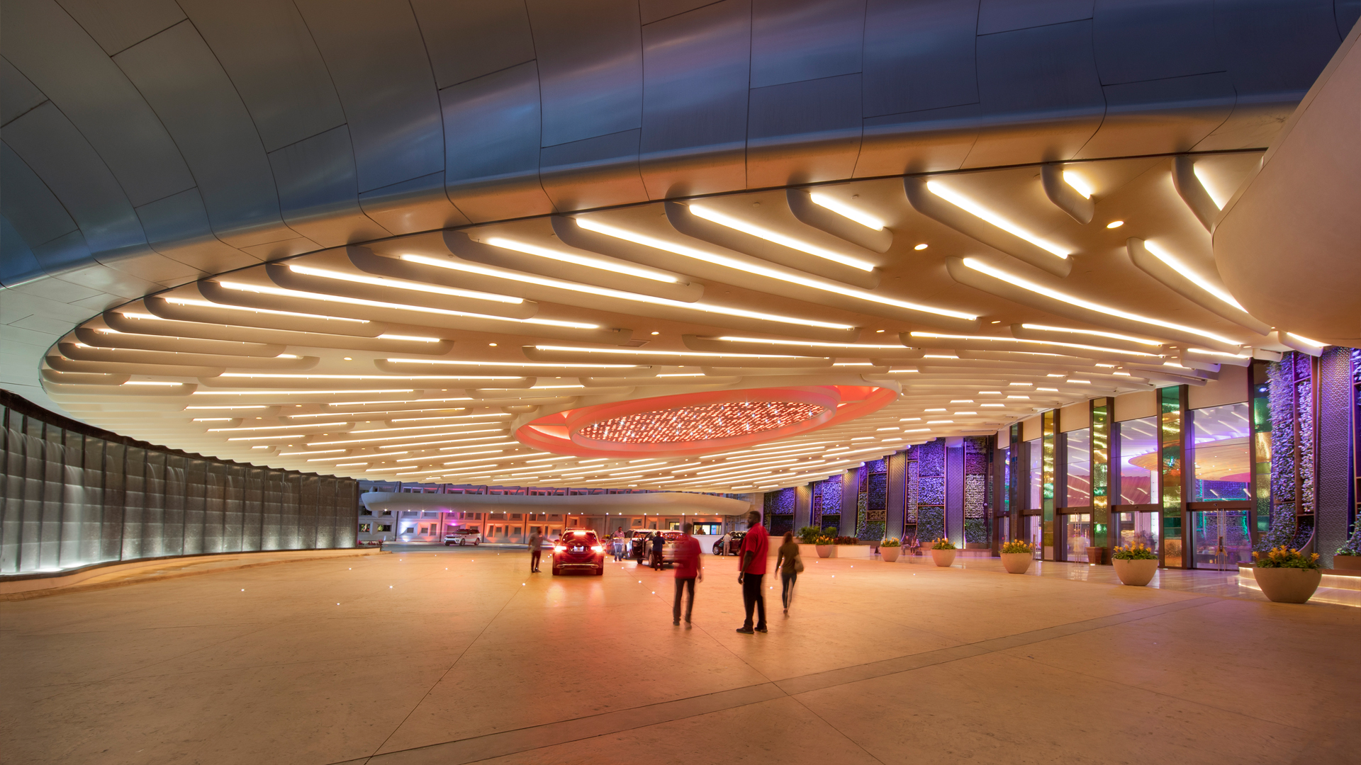People stand under a large, modern, brightly lit canopy with glowing patterns, outside a building entrance at night. Gaming enthusiasts gather nearby as colorful lights illuminate the façade and parked cars enhance the lively atmosphere.