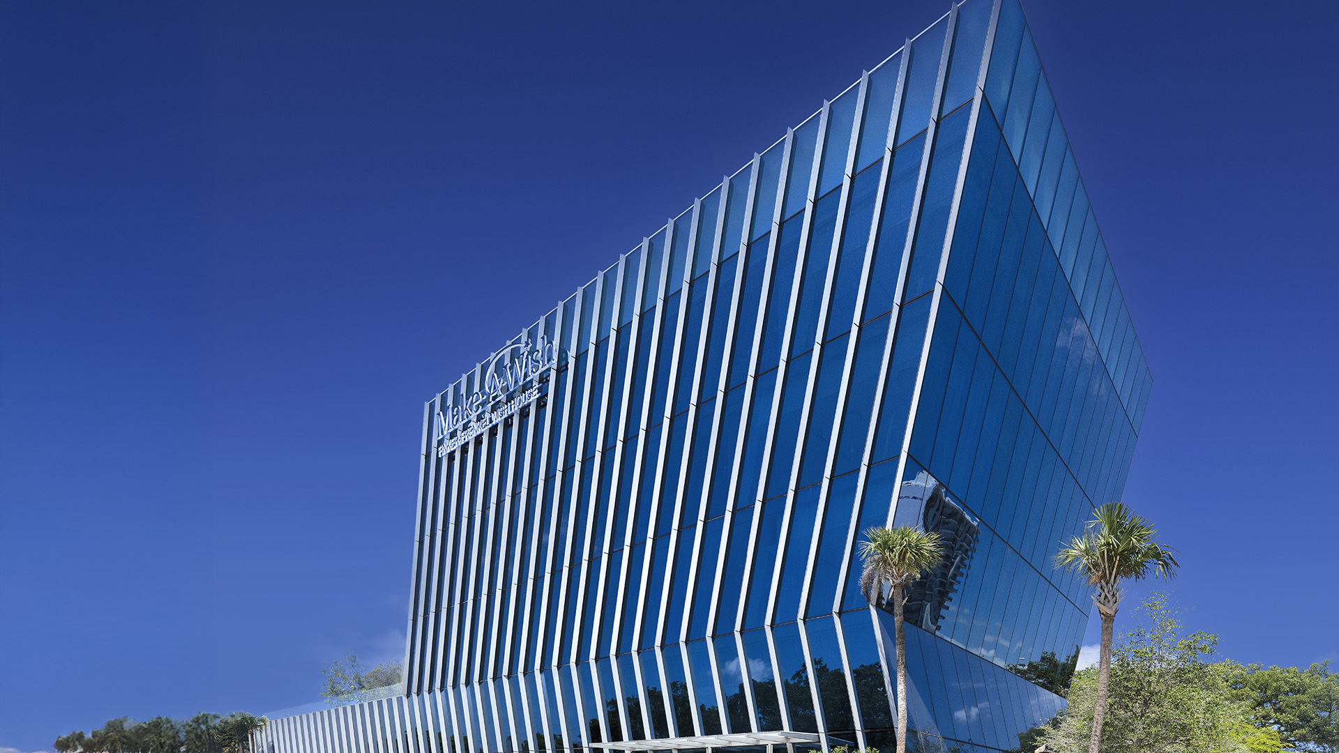 A modern glass office building with a slanted design reflects the blue sky. Palm trees line the front, and Nicklaus Children’s Hospital is displayed on the office building’s upper facade.