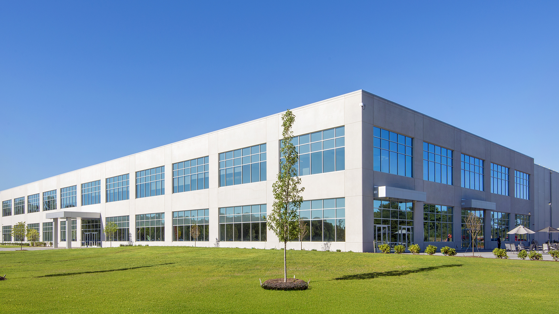 A modern, two-story distribution office building with large glass windows sits on a well-manicured lawn under a clear blue sky. There are a few small trees and outdoor seating with umbrellas near the entrance.