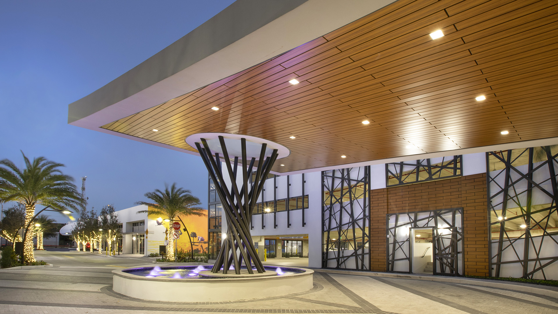 Modern affordable housing entrance at dusk with a large wooden overhang, unique black metal column, circular fountain, geometric window designs, palm trees, and illuminated lights creating a vibrant, welcoming atmosphere.