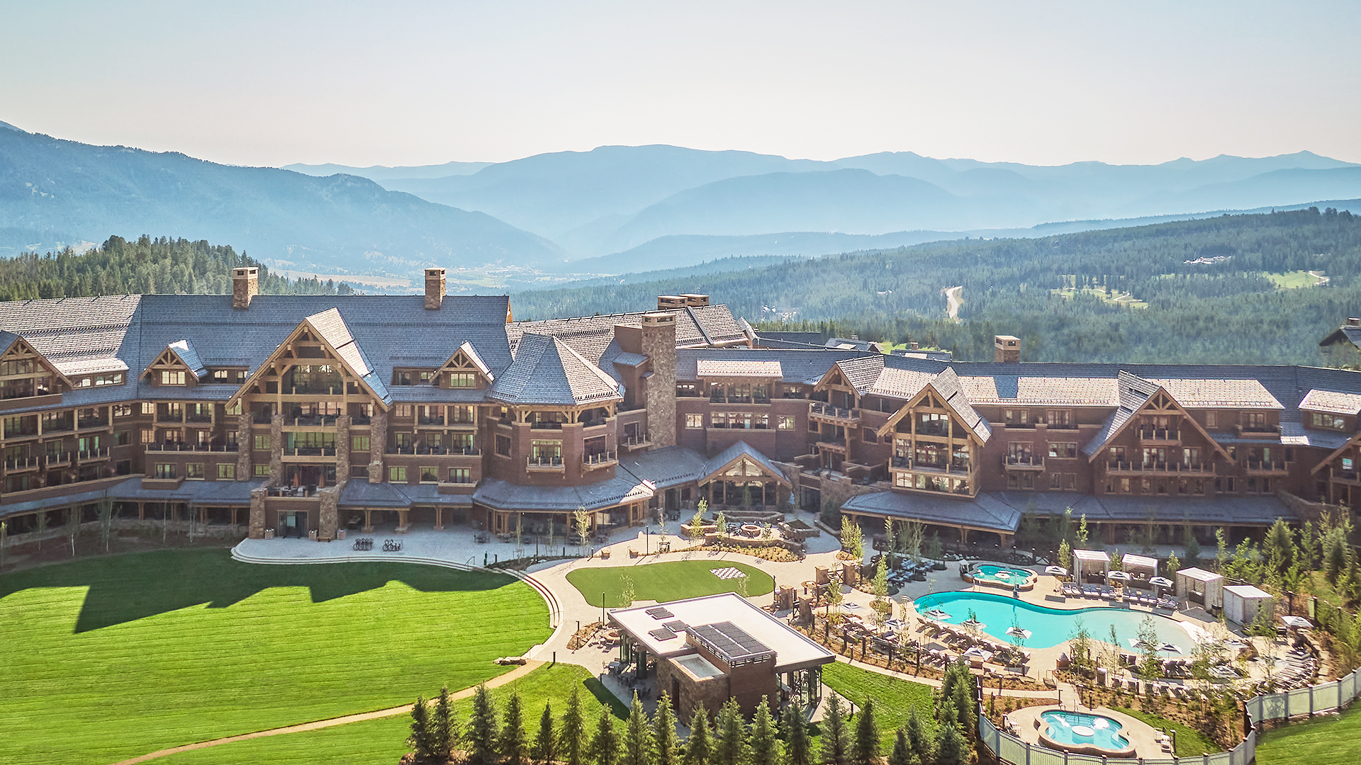 A large residential mountain resort with wood and stone architecture sits among green hills and trees. There is a spacious lawn, outdoor pools, and lounge areas, with forested mountains and a hazy sky in the background.