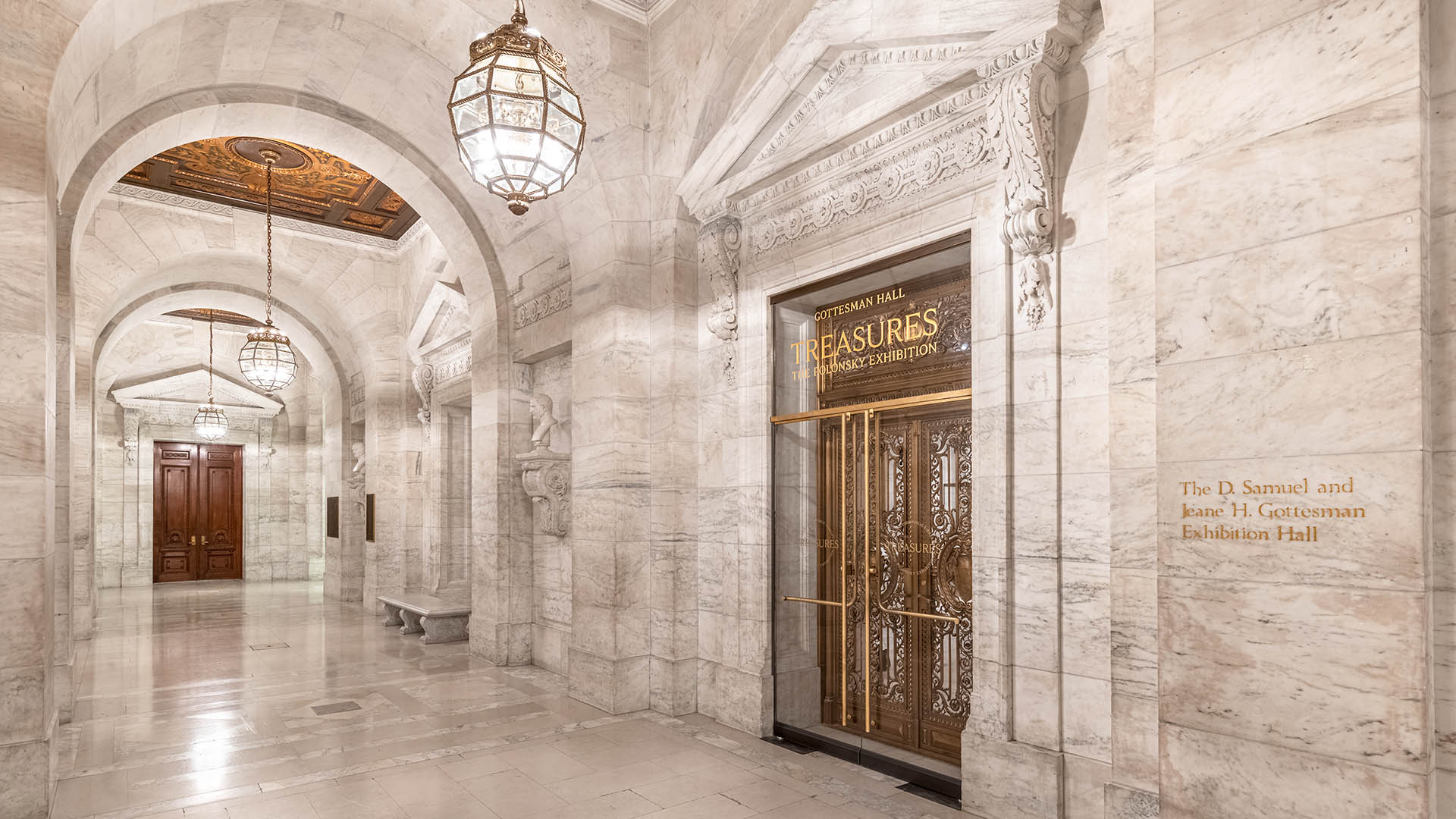 A grand marble hallway with ornate arches, decorative lighting fixtures, and gold-accented doors labeled “Treasures Exhibition.” The elegant, nonprofit venue is bright and welcoming, featuring intricate architectural details throughout.