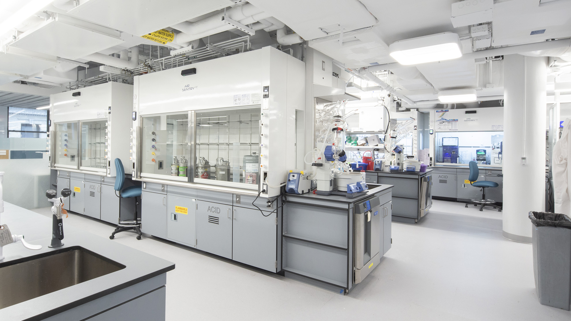 A modern, spacious Life Sciences laboratory featuring fume hoods, lab benches, blue chairs, and advanced scientific equipment. The well-lit space has white walls and ceiling, with various tools and containers on the workstations.