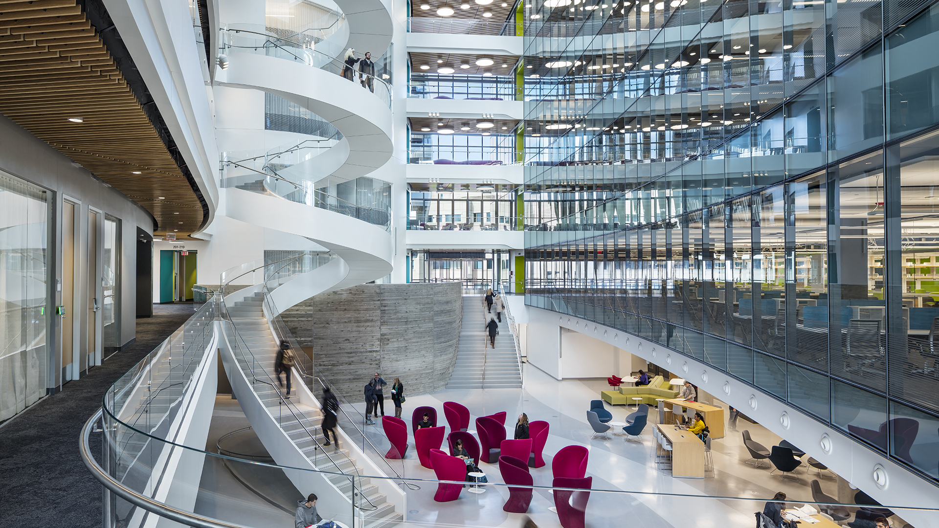A modern, spacious atrium with glass walls, a white spiral staircase, open walkways, and red lounge chairs creates an inviting space where people walk or sit in this bright, multi-story higher education office building.