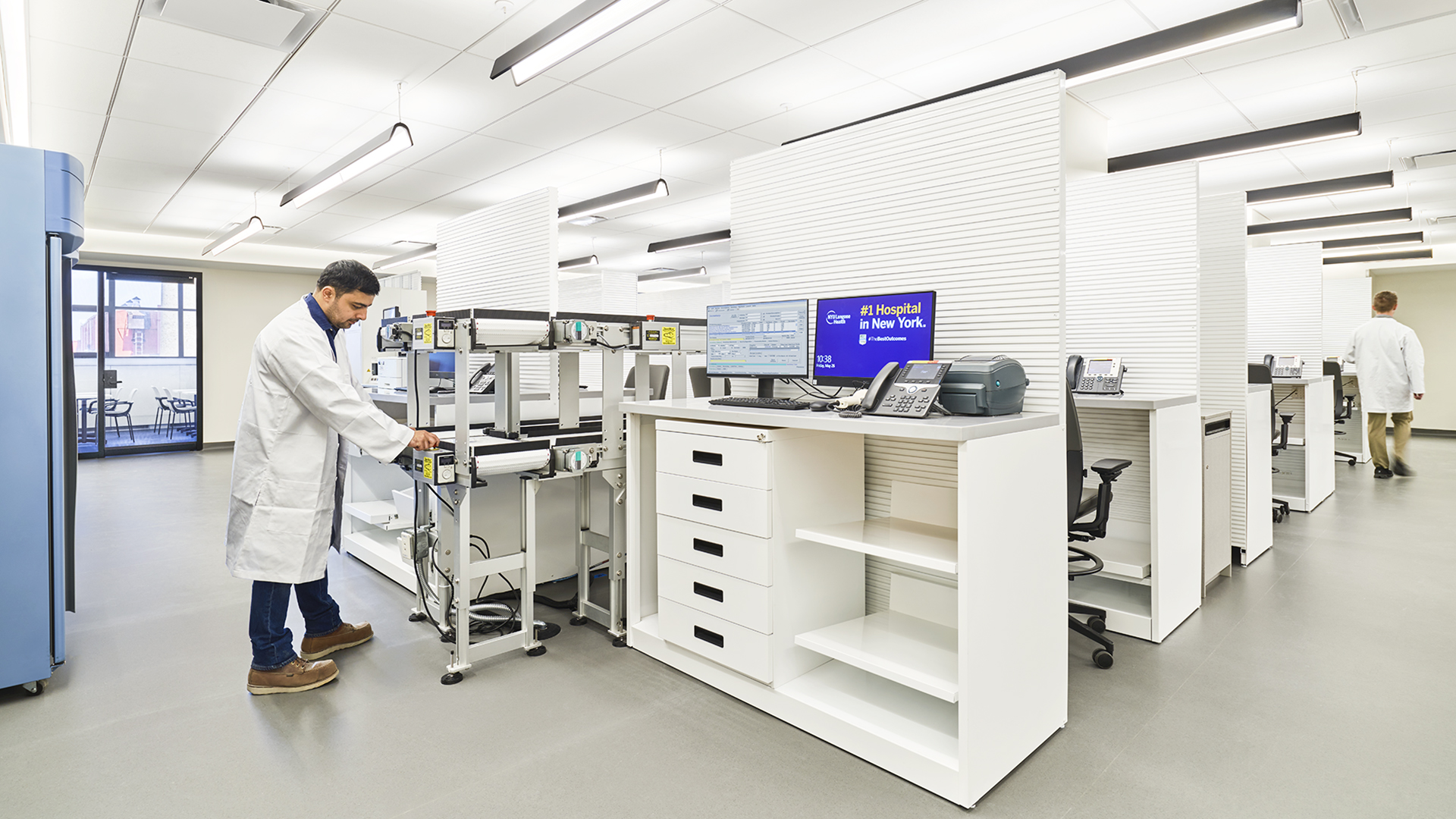 A man in a white lab coat works with equipment in a bright, modern healthcare laboratory. Computer monitors, shelves, and desks are visible; another person in a lab coat stands in the background. The space is clean and organized.