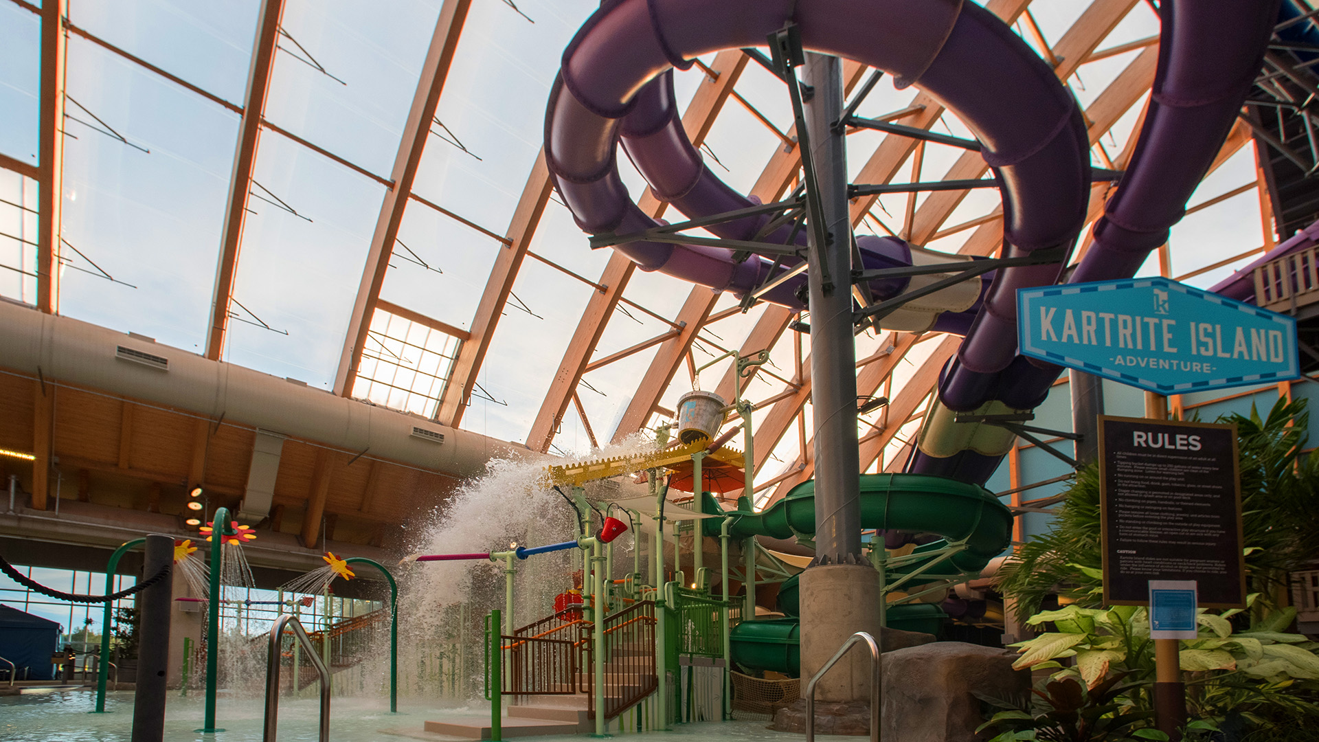 Indoor water park with colorful water slides, splash pads, and play structures under a high, curved glass roof offers nonstop entertainment. A sign reads Kartrite Island Adventure near a black rules board.
