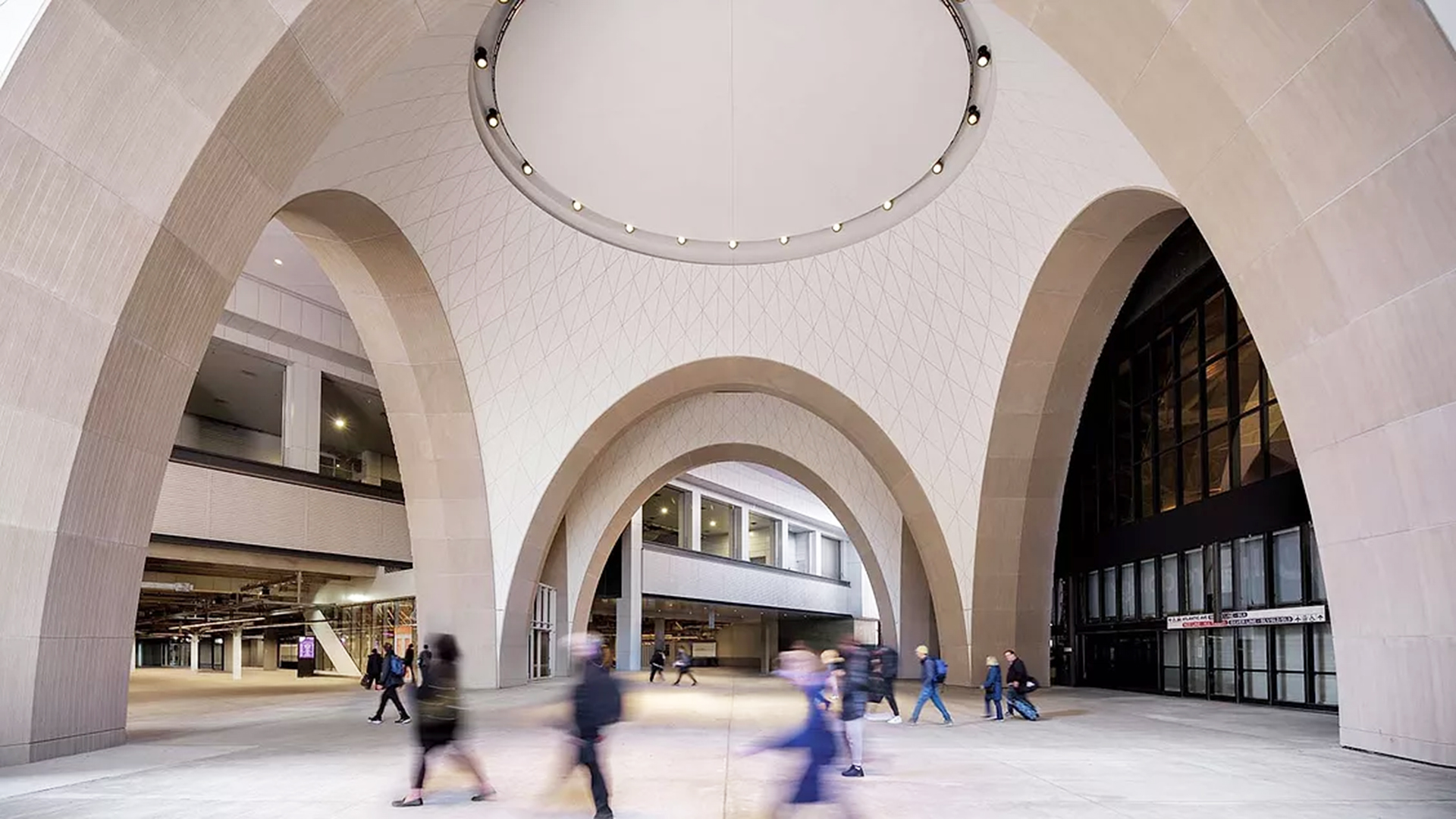 People walk under large, modern concrete arches in an open plaza area outside an office complex, with blurred motion suggesting movement. The space is surrounded by buildings and features a circular ceiling with lights.
