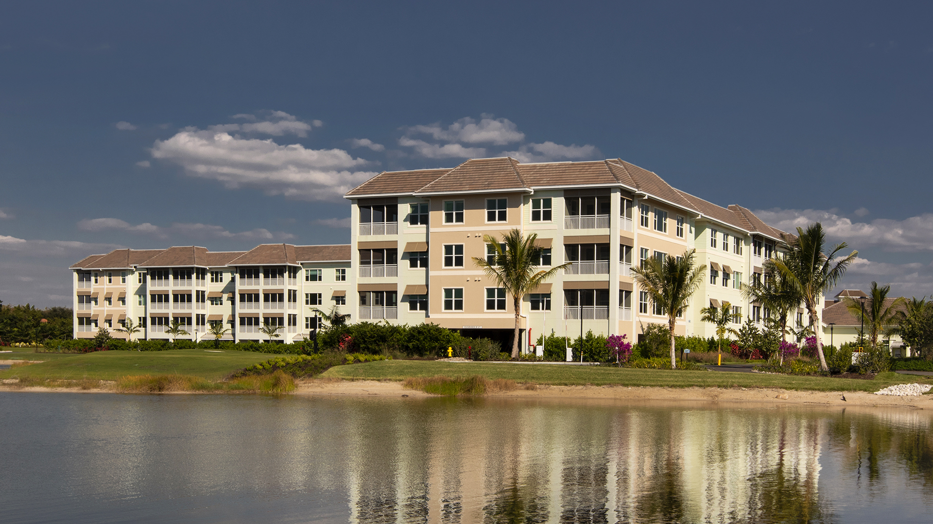 Three modern, light-colored assisted living apartment buildings with balconies are surrounded by palm trees, facing a calm pond under a clear blue sky with scattered clouds. A manicured lawn runs along the water’s edge.