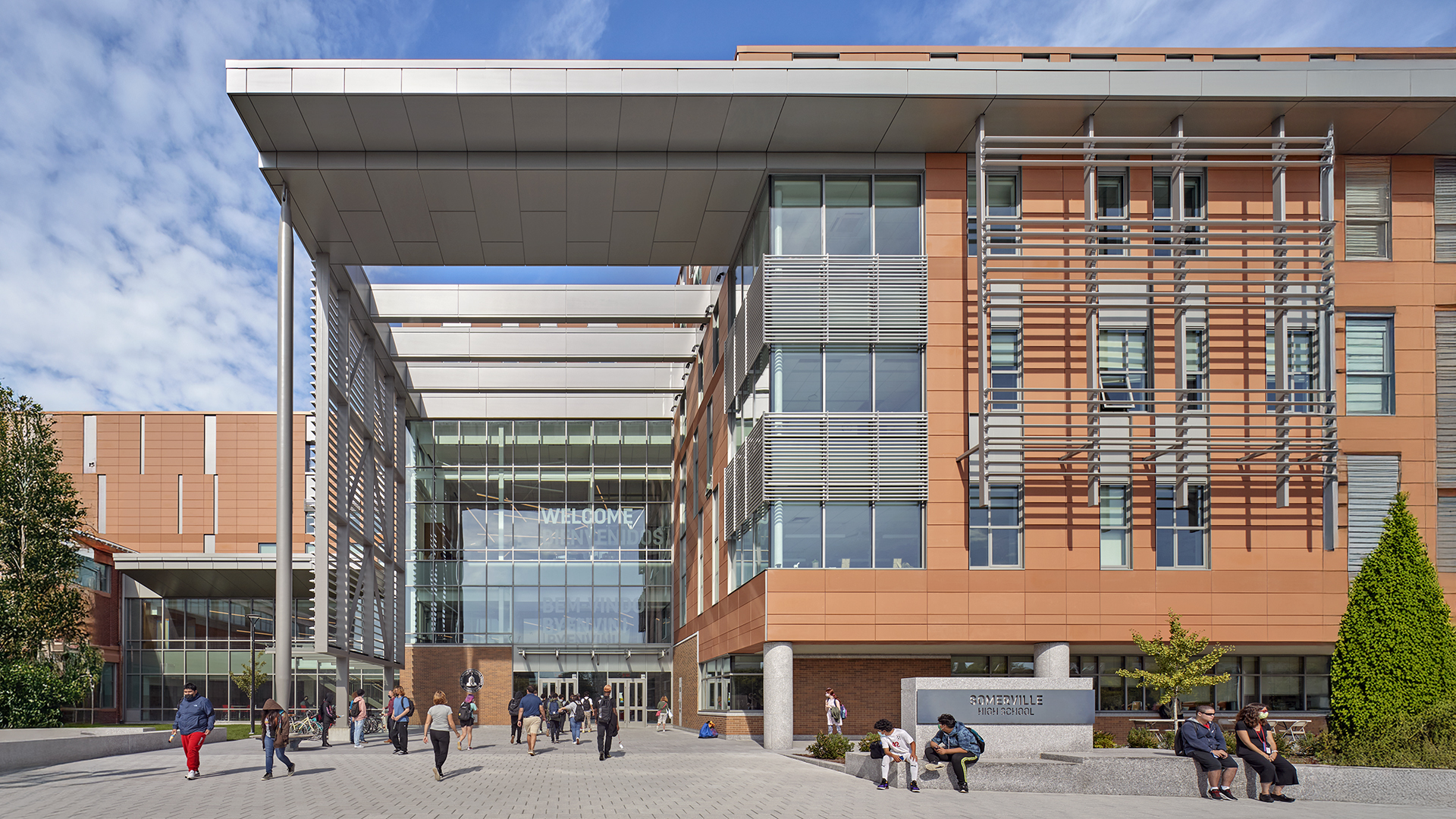 A modern university building with large windows, orange and gray panels, and a covered entrance welcomes both K-12 visitors and students. People walk and gather outside on a sunny day; “WELCOME” is visible above the main doors.