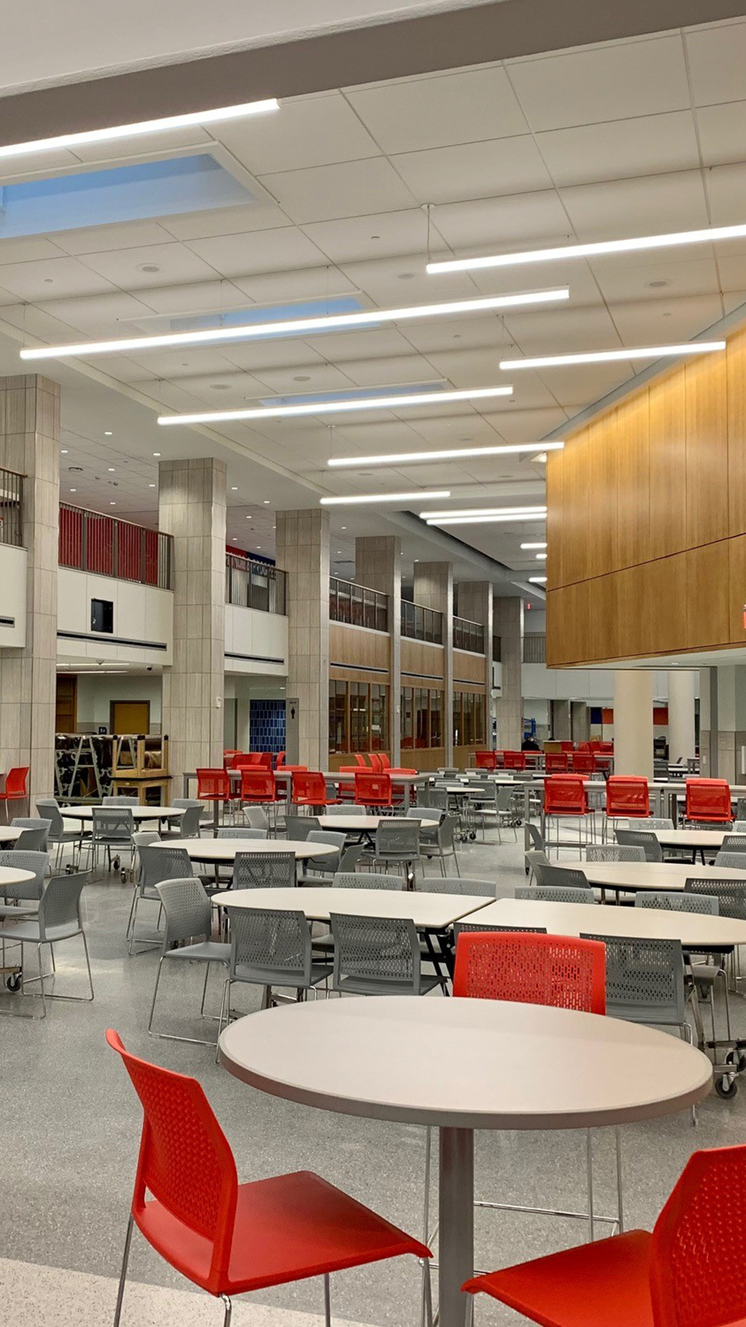 A spacious, modern cafeteria designed for K-12 students features round tables and red and gray chairs. The well-lit area includes tall pillars, large windows along the walls, and has only a few people present.