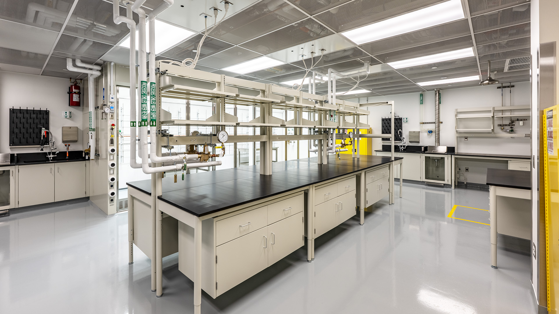 A modern, empty laboratory for Life Sciences with clean white floors, lab benches with black countertops, overhead shelves, safety equipment, and bright fluorescent lighting. No people are present.