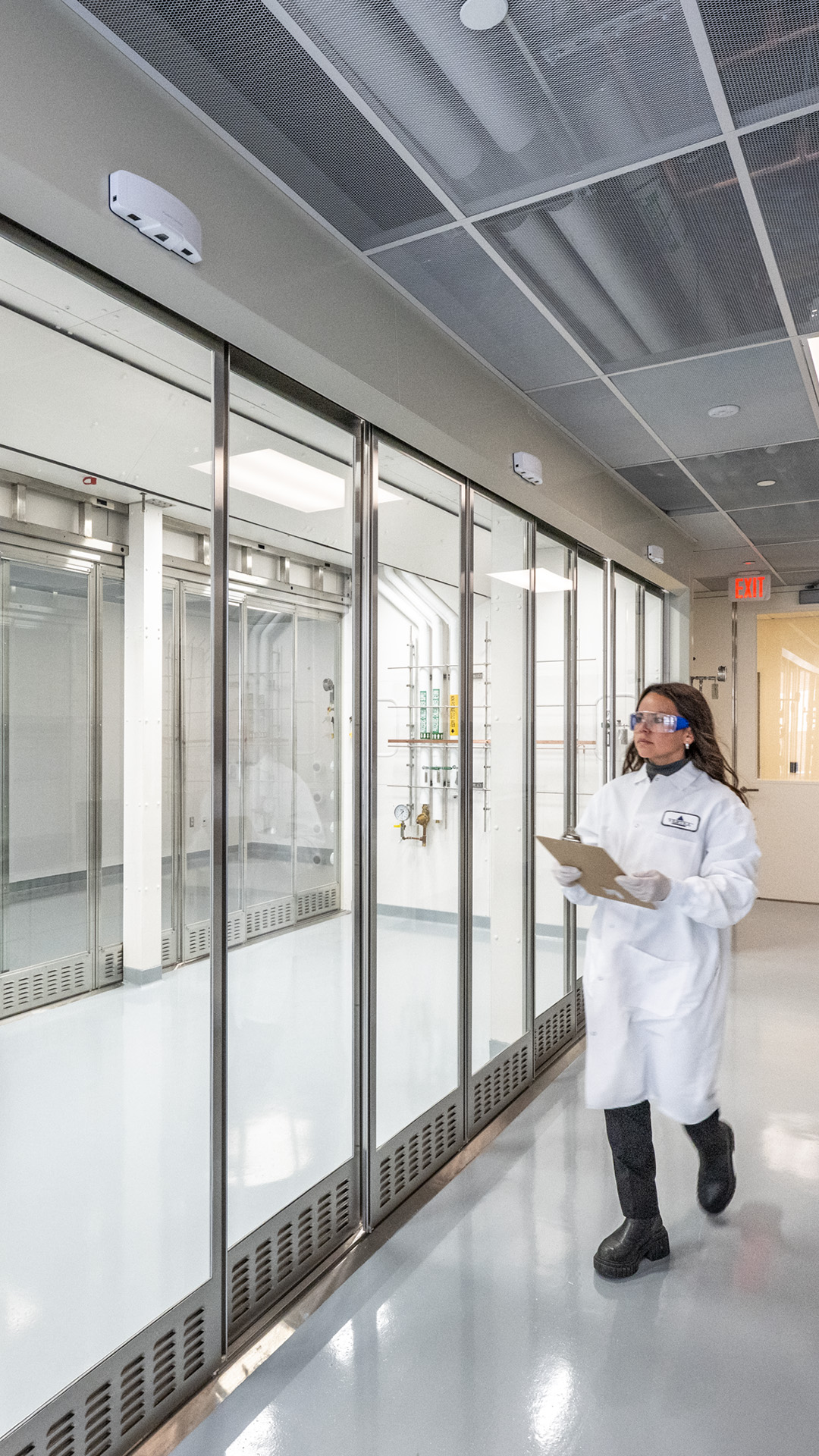 A person wearing a white lab coat, gloves, goggles, and boots walks through a bright, modern Life Sciences laboratory hallway, holding a clipboard. Large glass-walled rooms and scientific equipment are visible in the background.