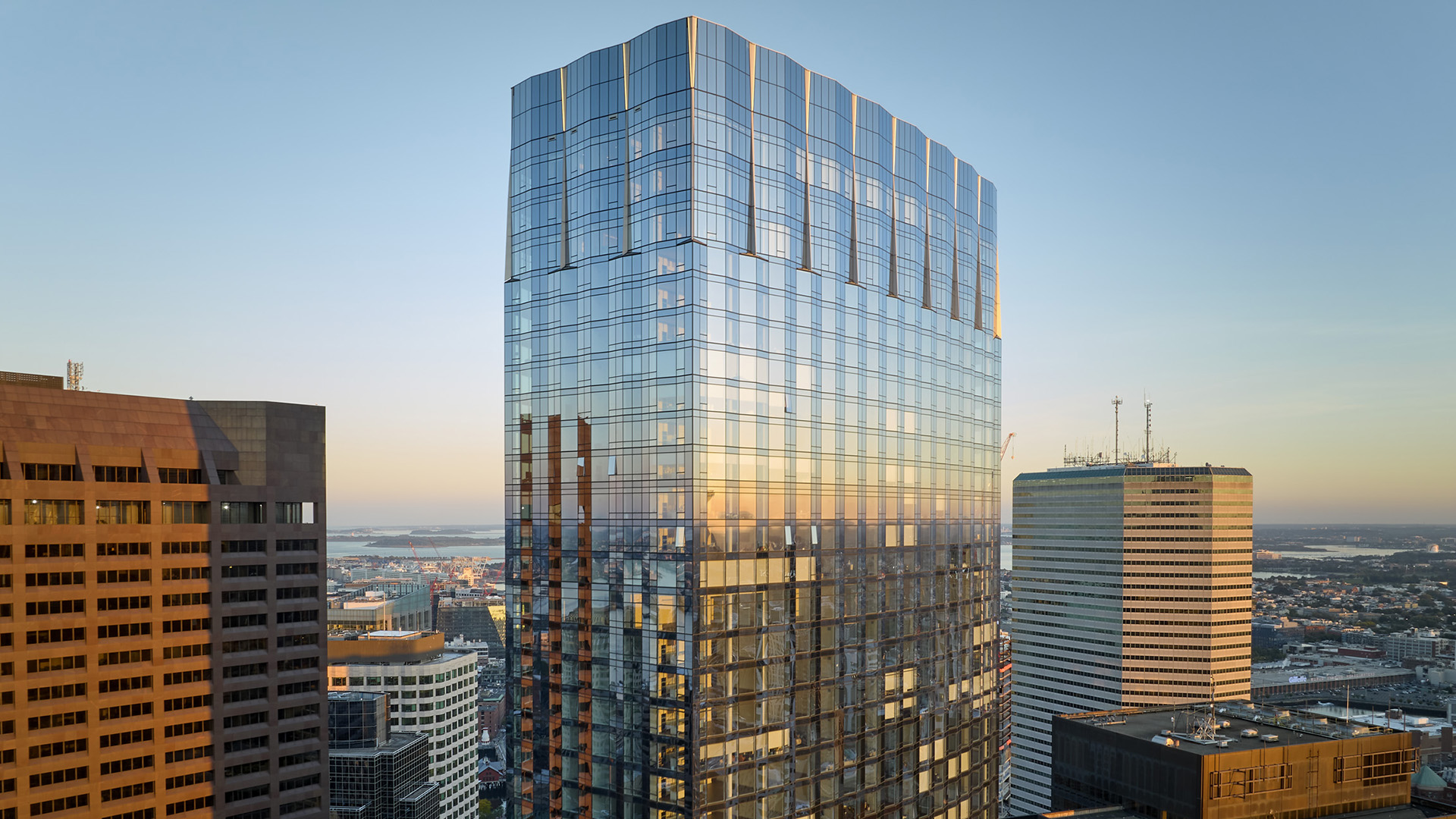 A modern glass office skyscraper reflecting the city skyline and sunset, surrounded by other tall buildings in an urban setting. The sky is clear and the cityscape extends into the distance.