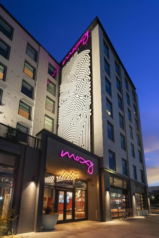 Modern hotel exterior at dusk with moxy neon sign above the entrance, abstract black and white mural on the building facade, and large windows reflecting the evening sky.
