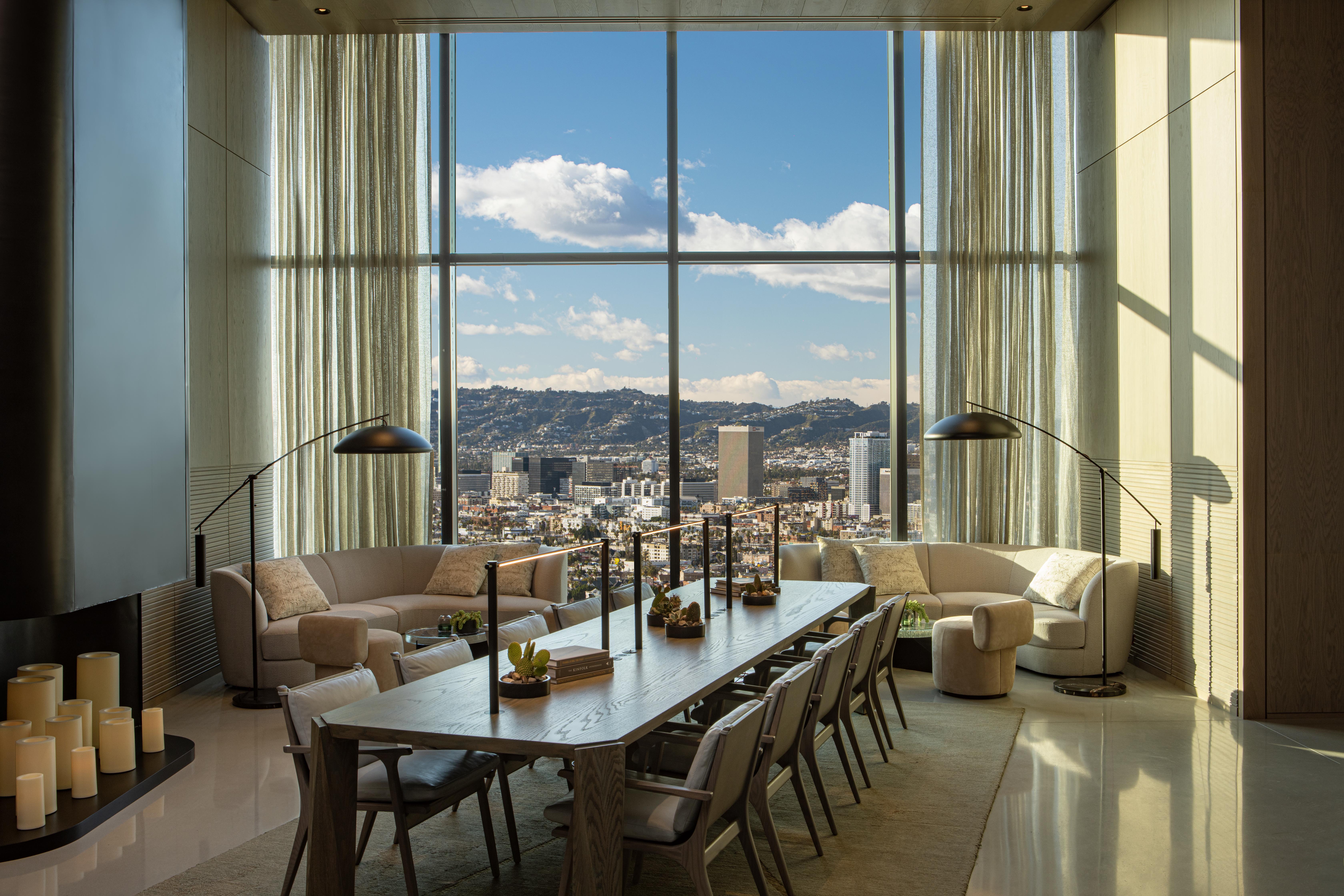 A modern dining room with a long wooden table, stylish chairs, and cozy seating areas by tall windows overlooking a cityscape and distant hills under a blue sky with clouds.
