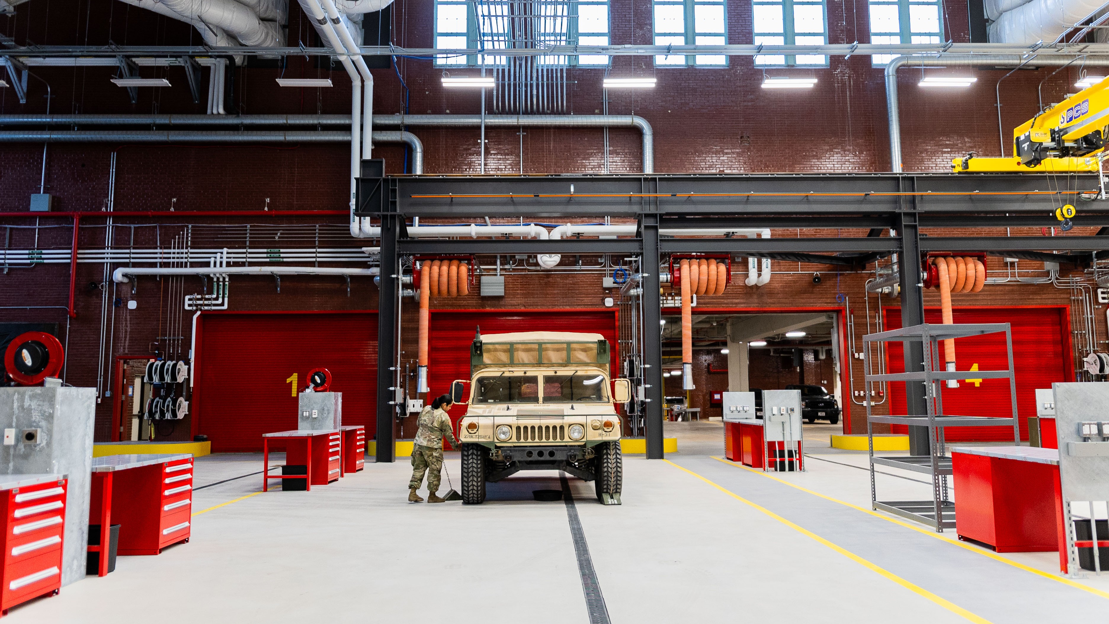 Two people in military uniforms stand next to a tan military vehicle inside a spacious, well-lit garage with red tool cabinets, workstations, and industrial pipes along the brick walls.
