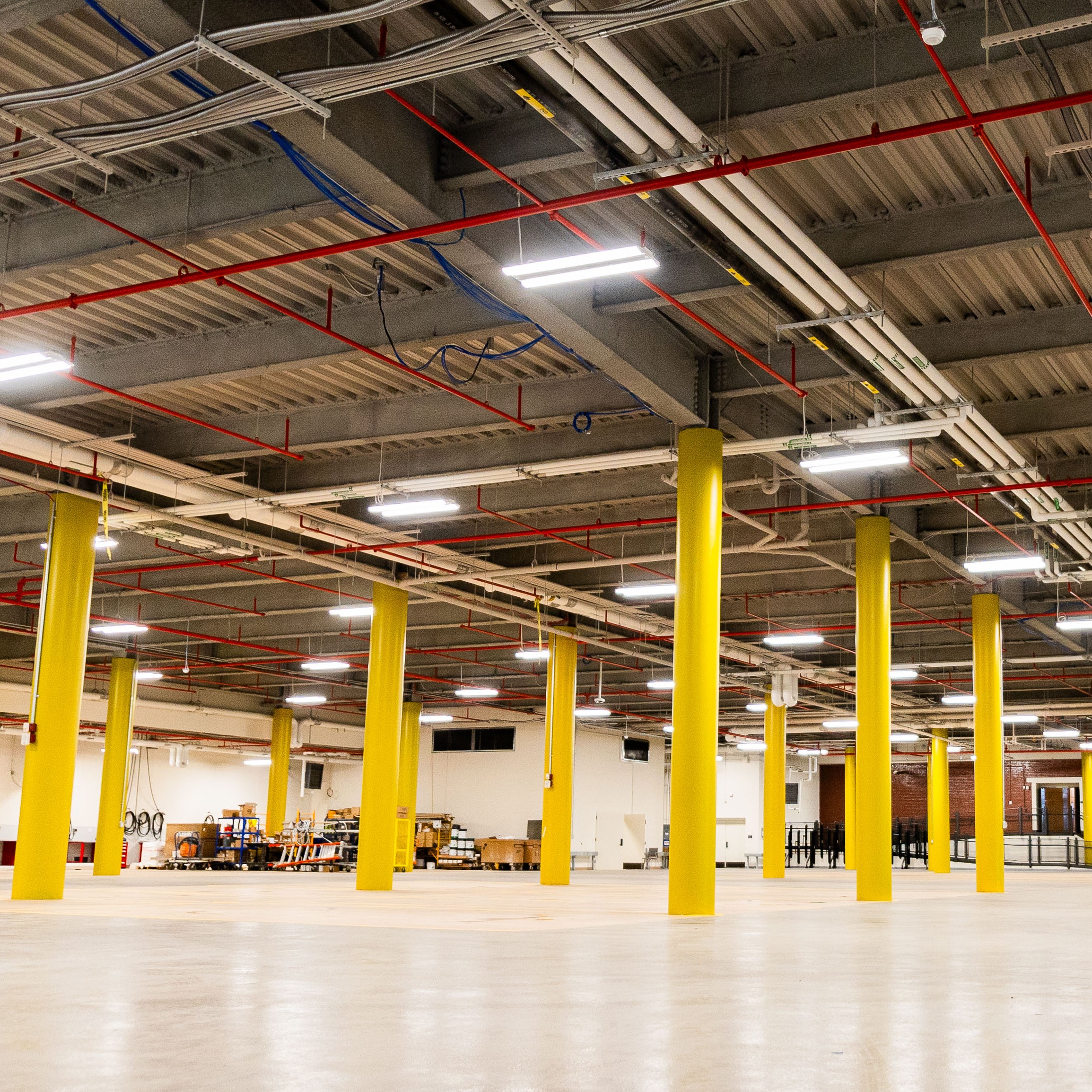 A large, empty indoor warehouse with yellow support columns, exposed ceiling pipes, and scattered boxes and equipment along the far wall under bright fluorescent lighting.